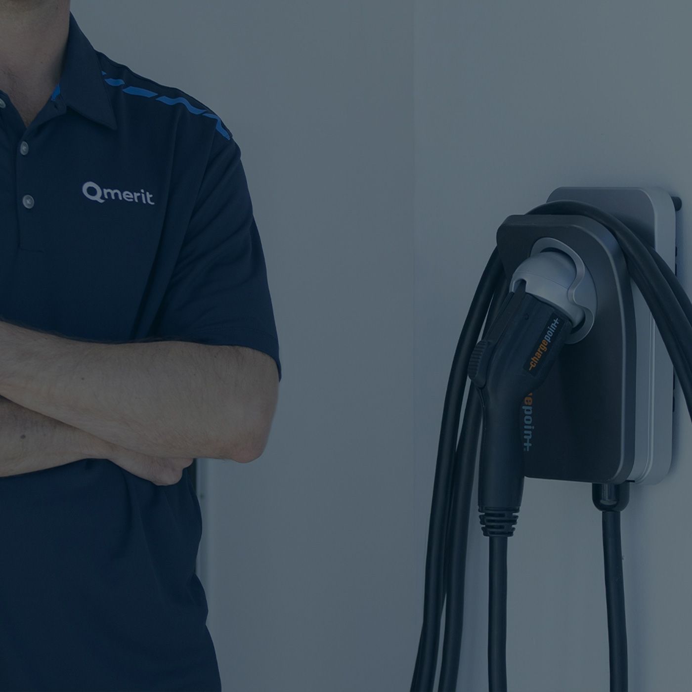 A two-shot of a person, with head cropped out, wearing a blue Qmerit uniform polo and crossing their arms in confidence, standing next to an installed home charger, including wall mount, charger, and long cable coiled around the mount.