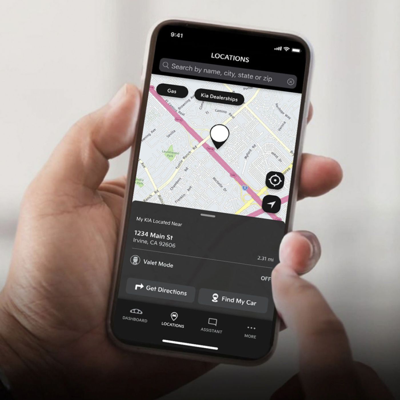 A hand holding a mobile phone, on which is displayed a map and a place marker showing the location of the user's parked Kia vehicle, allowing them to find their car. Options for gas, Kia Dealerships, Valet Mode, Find My Car, Assistant, and more also appear on the screen.