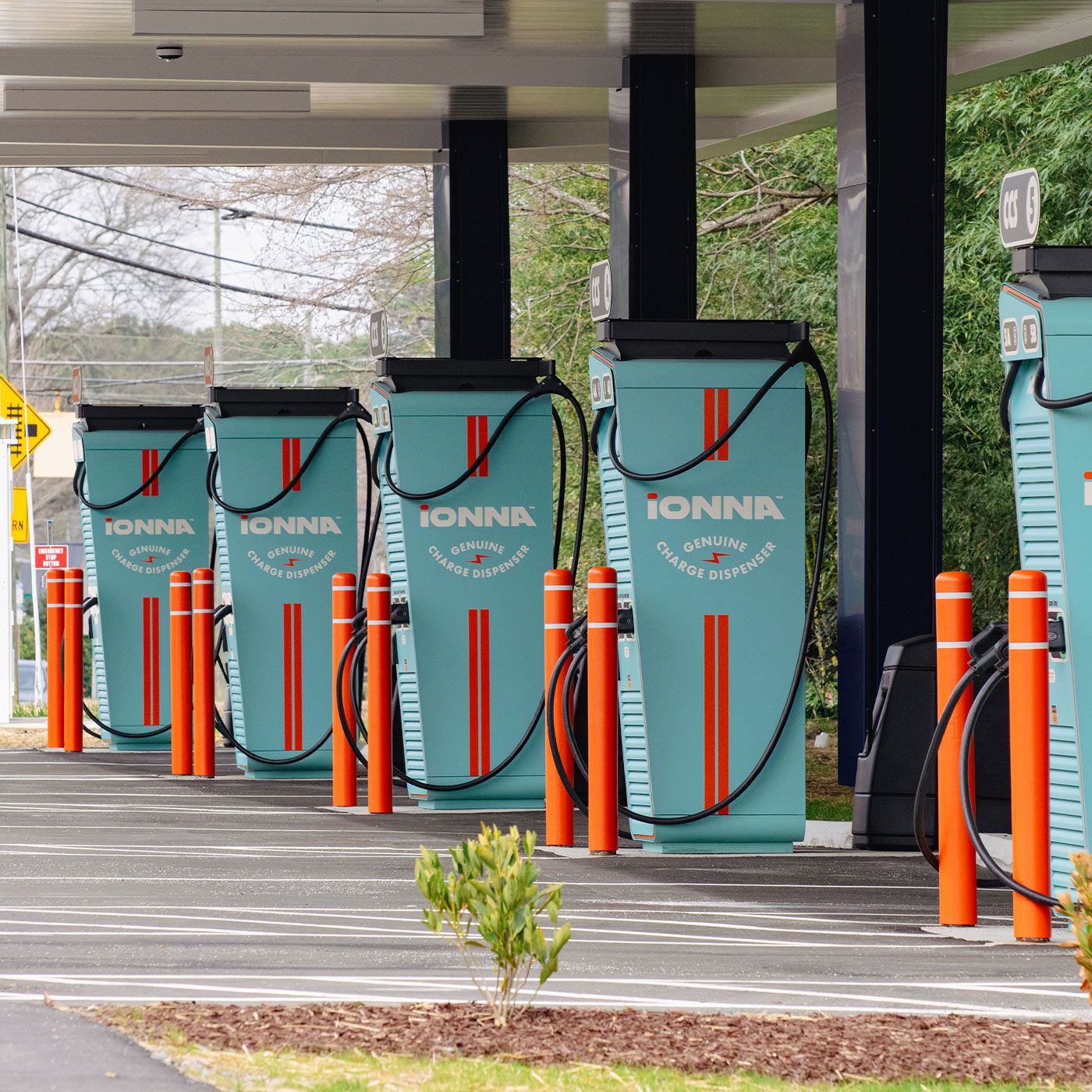 A row of pastel blue iONNA hybrid-electric vehicle charging stations, with bright red stripes, black charging cables, and orange safety beams, under a shelter with trees and fixtures in the background. The spaces are all open and ready for a Kia vehicle to use.