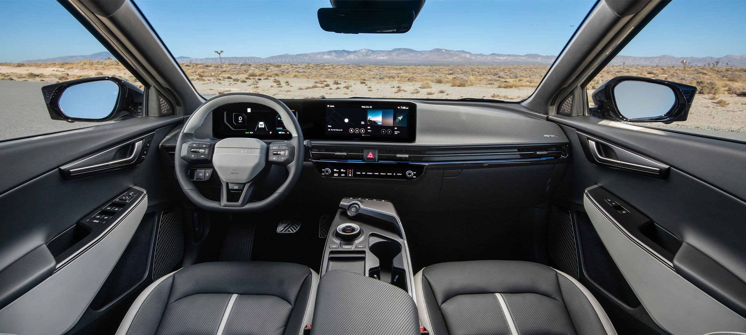 An interior front view of the 2026 Kia EV6, with a desert view and distant mountain range outside of the windows and windshield. The car's interior features a steering wheel, 12.3 inch dual panoramic displays, the dashboard, rearview mirrors, and more.