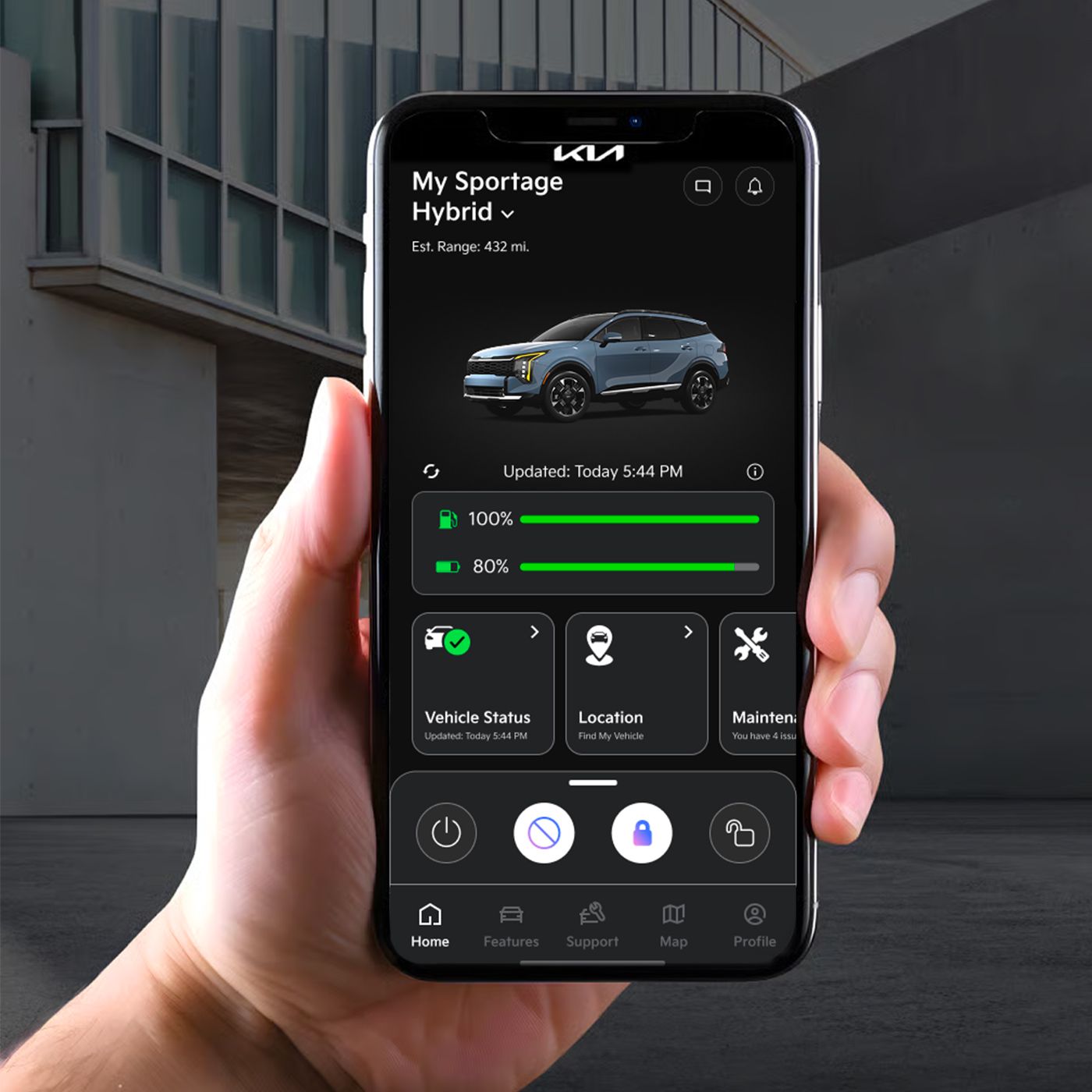 Hand holding a smartphone displaying the Kia vehicle app with a Kia SUV shown at 100% fuel with features to check vehicle status, location, and more, against an architectural background.