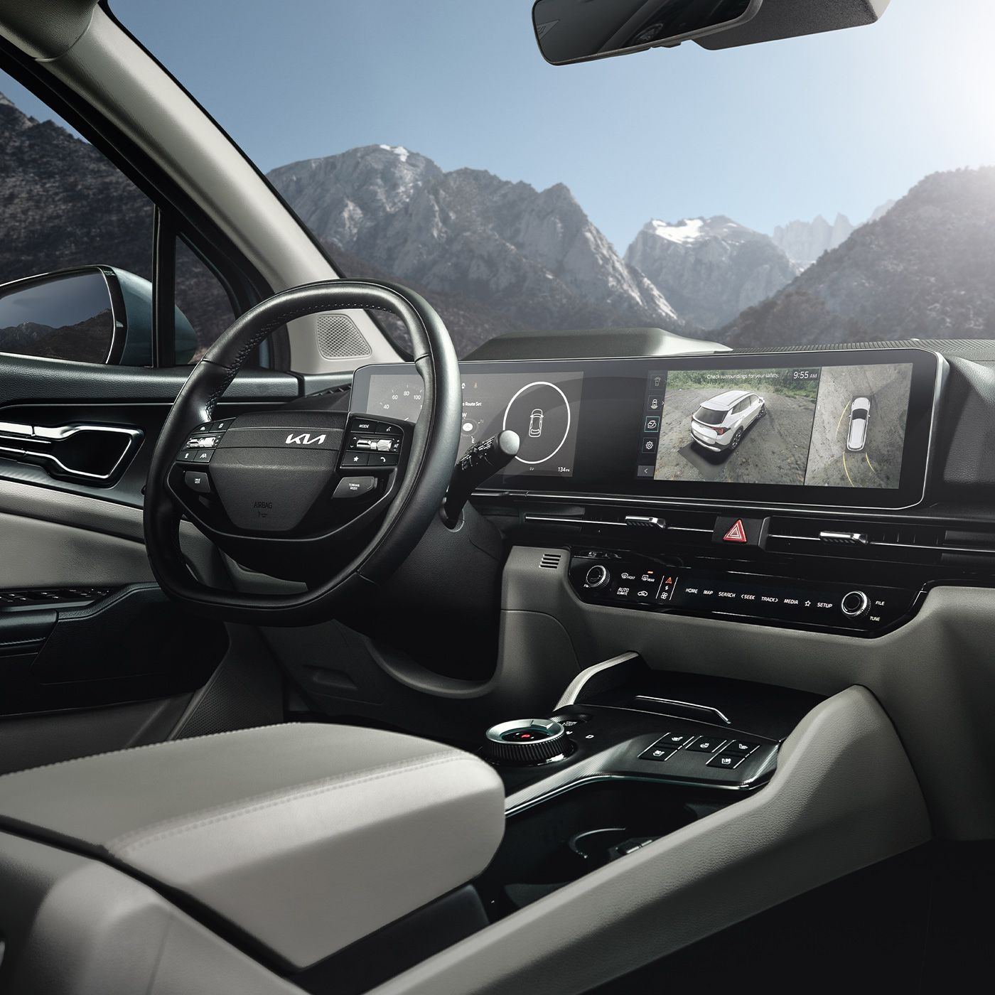 Interior view of the 2027 Kia Sportage Hybrid in gray featuring the driver-side steering wheel, dual panoramic display, and the center console with scenic snowy mountains and blue skies visible through the windshield.