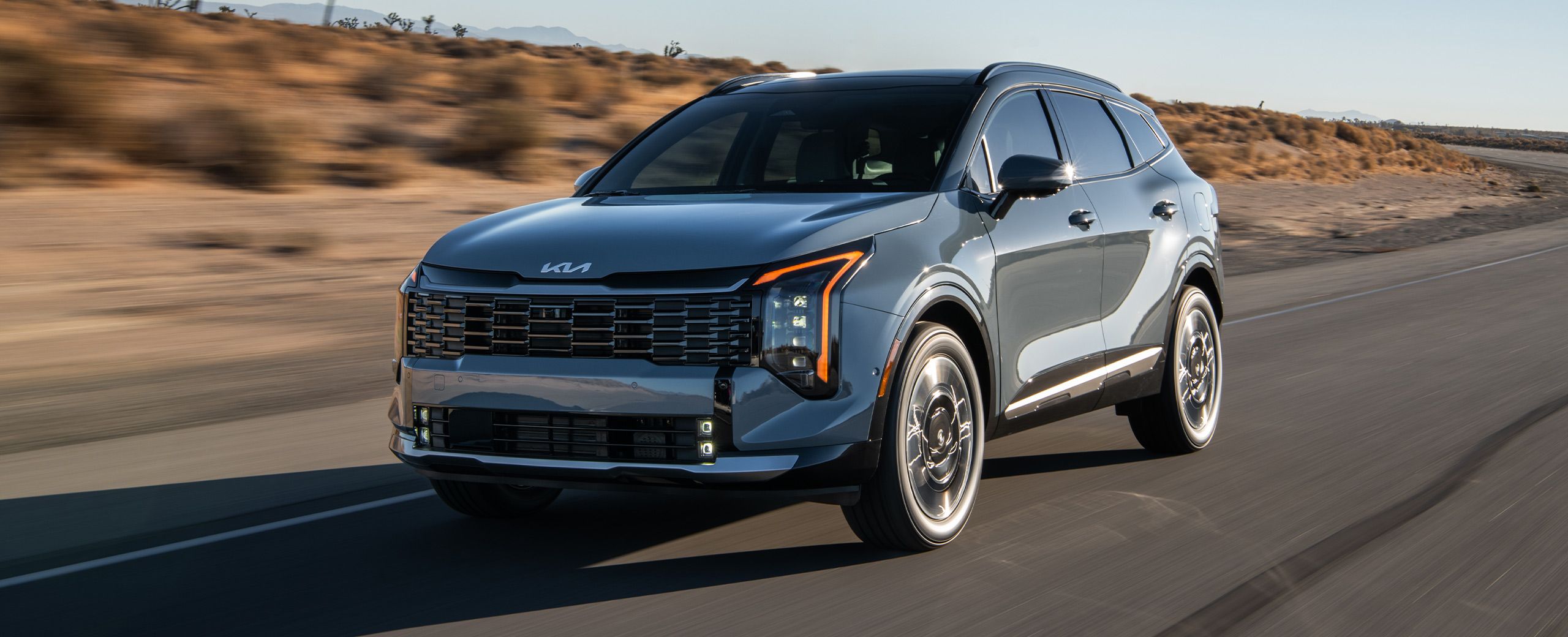 2027 Kia Sportage Hybrid driving on a desert highway, shown in a front three‑quarter view with vertical LED headlights, sculpted body lines, and distinctive wheel styling.