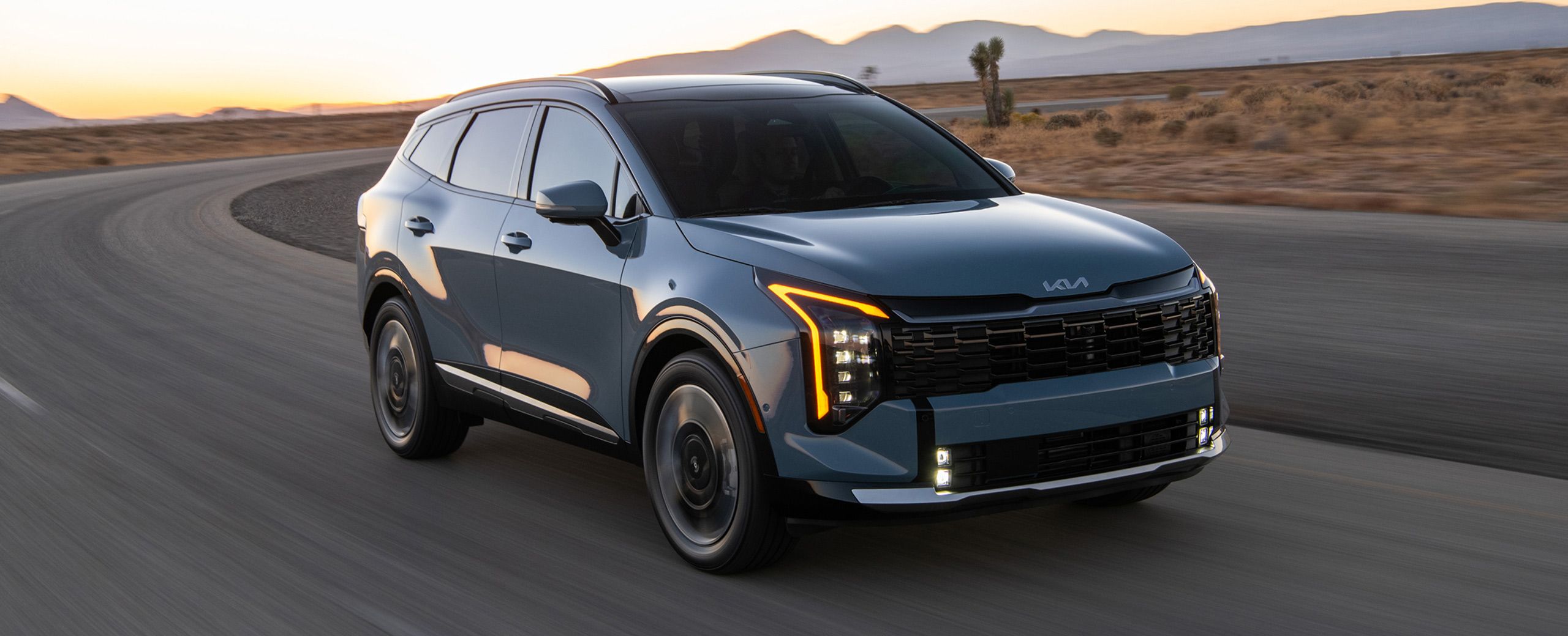 2027 Kia Sportage Hybrid driving on a winding desert highway with mountains in the background during sunset, shown in a front three‑quarter view with vertical LED headlights and amber daytime running lights being highlighted.
