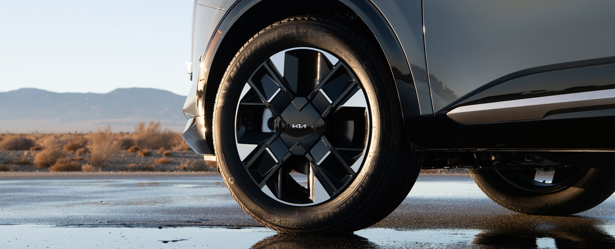 A close-up shot of the 2027 Kia Sportage Hybrid 19-in. distinctive front wheels parked on a wet road with mountains and plants in the background.