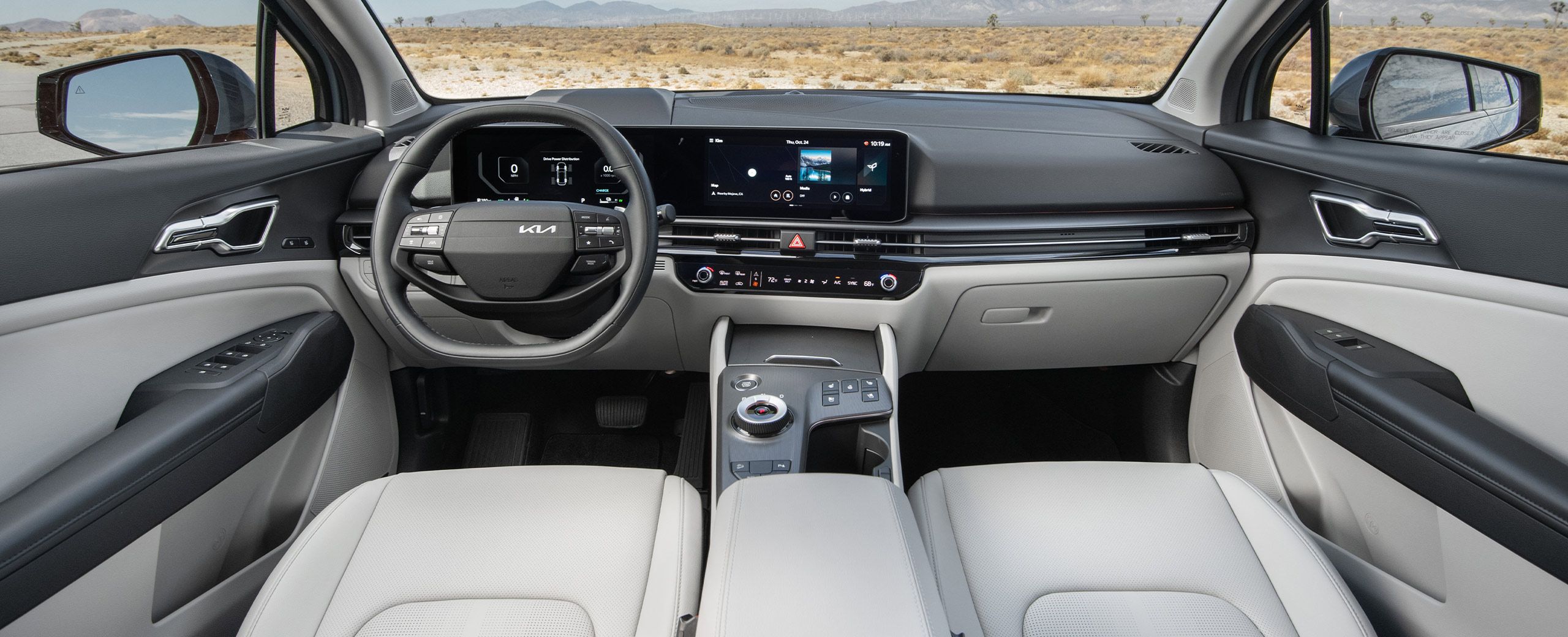 Interior view of the 2027 Kia Sportage Hybrid featuring a light gray interior with the driver-side steering wheel, digital display, sleek design, and center console highlighted, with a desert landscape and distant mountains visible through the windshield.