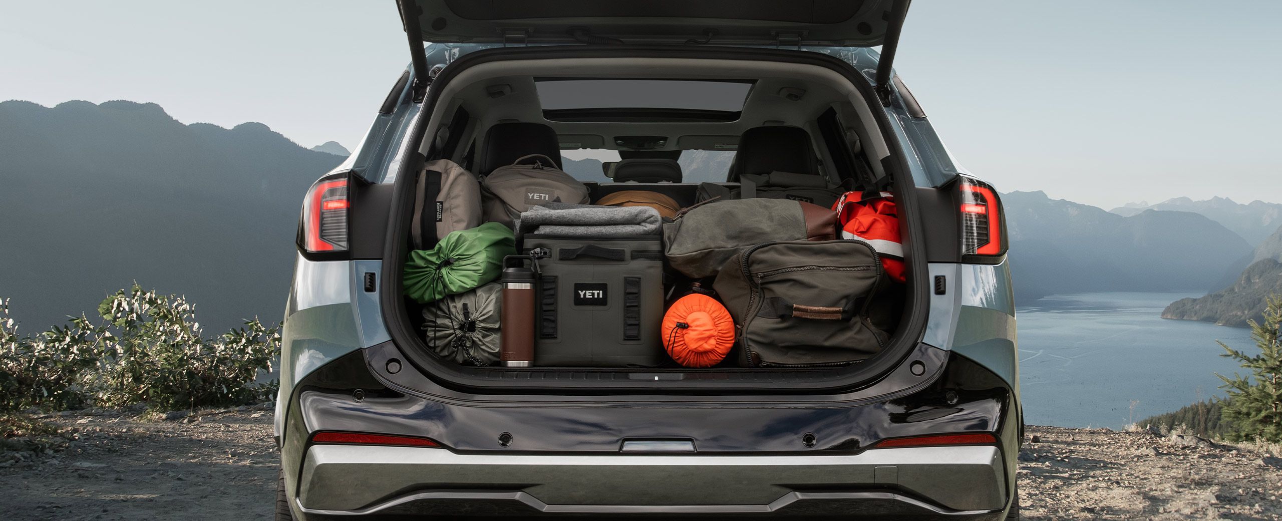 Photo of the 2027 Kia Sportage Hybrid with an open trunk showcasing the spacious cargo area packed with outdoor gear, parked against a cliff with mountains and a lake visible.