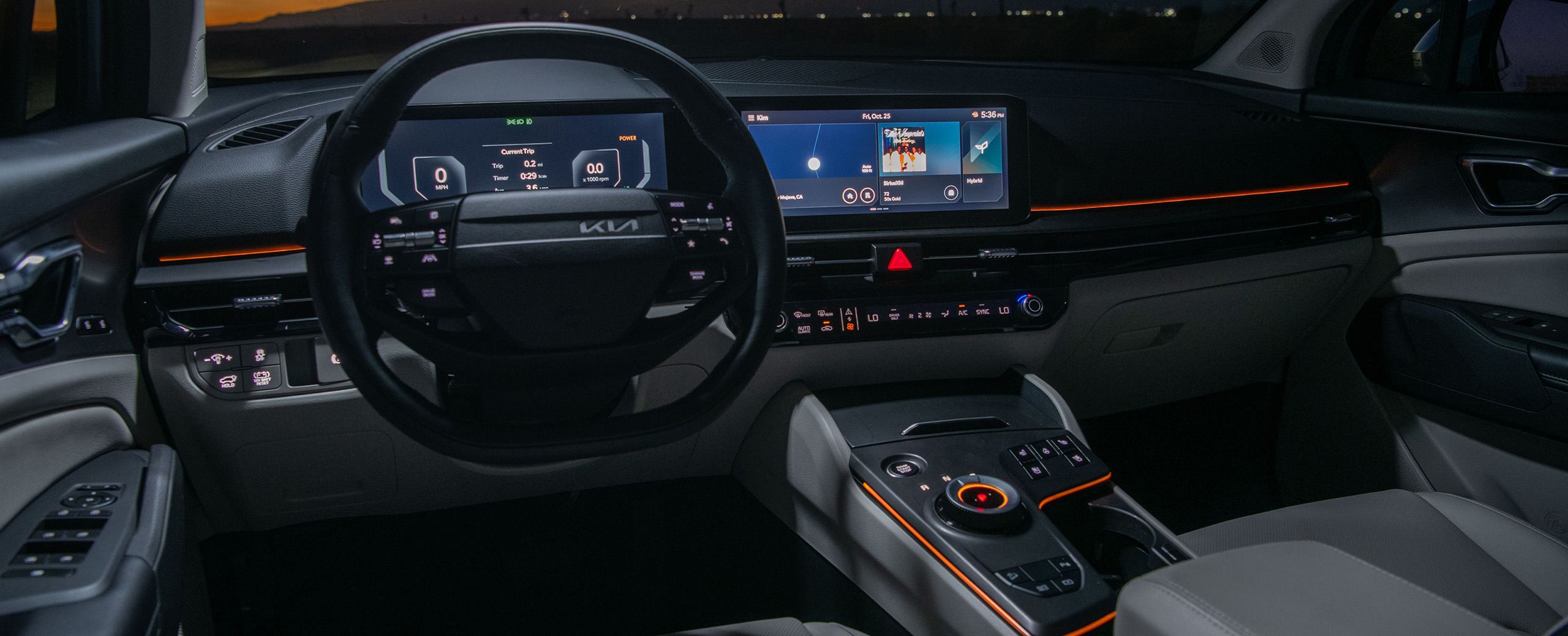 Interior view of the 2027 Kia Sportage Hybrid cockpit with a digital instrument cluster, wide infotainment display, and color‑selectable ambient mood lighting set to red, illuminating the dashboard and center console with a sunset and mountains visible through the windshield.