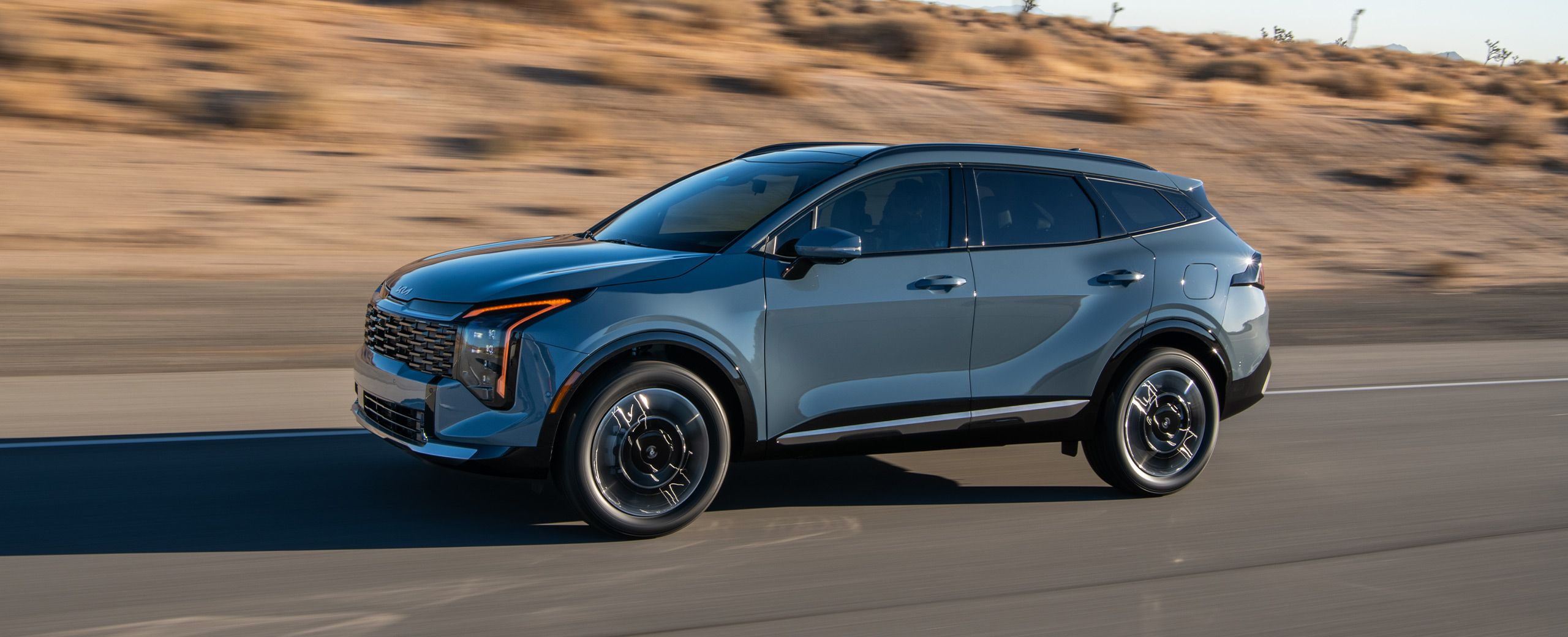 A blue 2027 Kia Sportage Hybrid compact SUV is driving along a desert highway, highlighting the driver's side and sleek design.