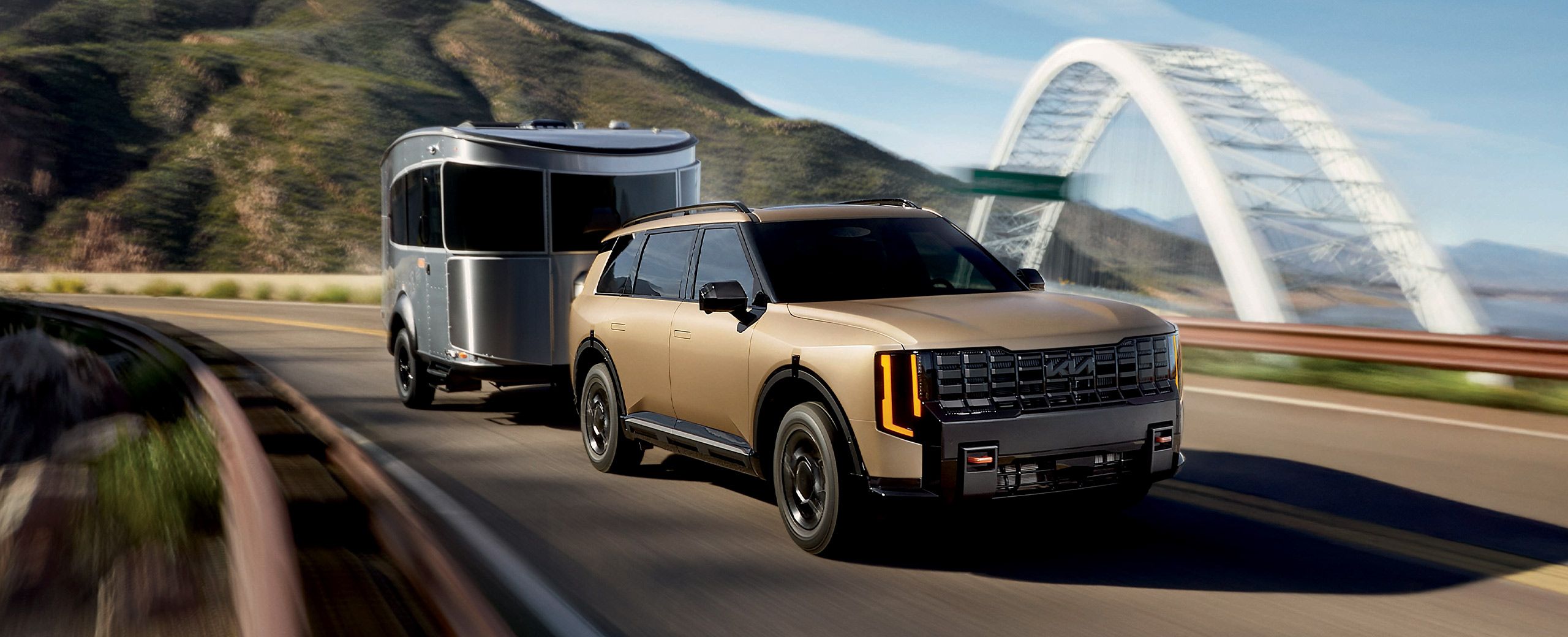 Tan Kia Telluride towing a silver travel trailer on a curving road, with an arched bridge and hills behind