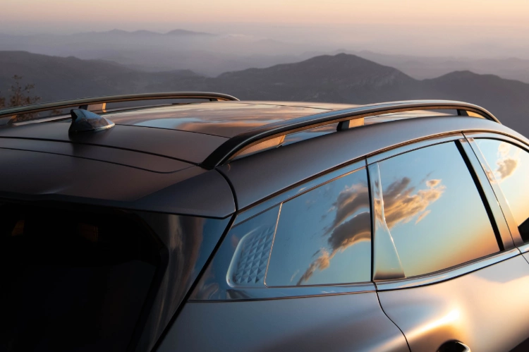 2024 Kia Sportage Plug-in Hybrid in matte gray in a three-quarter side view of the roof, featuring roof rails and darkened windows that reflect sunset colored clouds