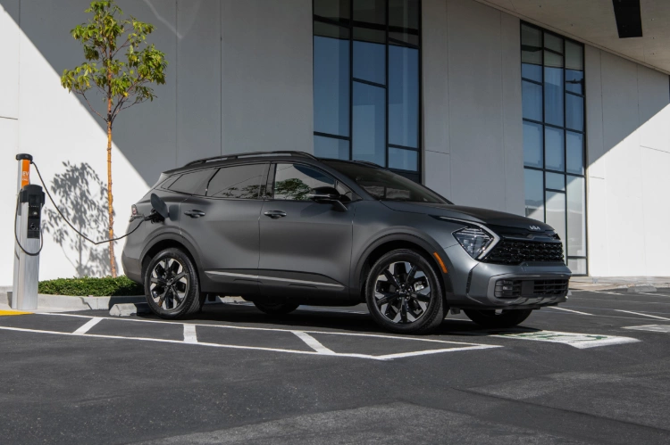 2024 Kia Sportage Plug-in Hybrid, matte gray in a three-quarter front view, plugged into a charging station while parked in front of a building