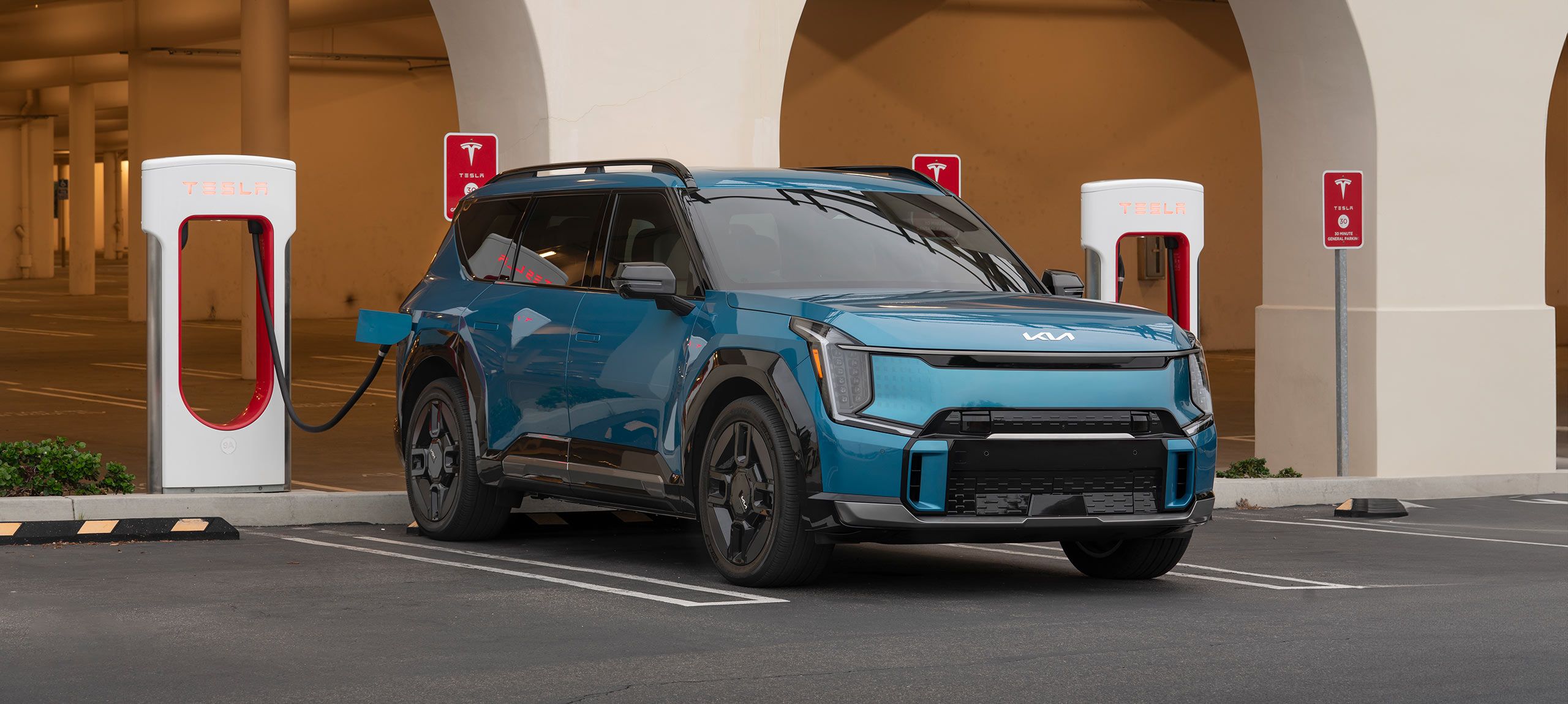 A blue Kia EV9 parked at a Tesla Supercharger station, shown for illustration