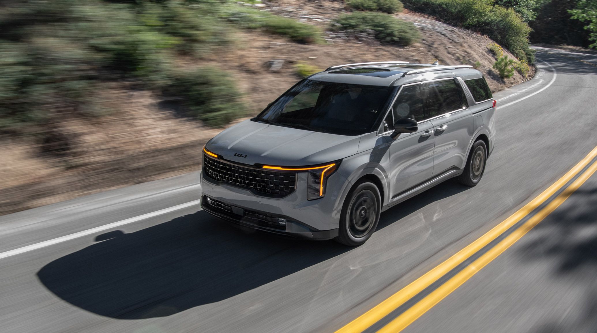 2027 Kia Carnival Hybrid Minivan driving along a winding forest road, highlighting its roof rails and dual sunroofs with the light shining off the gray exterior.
