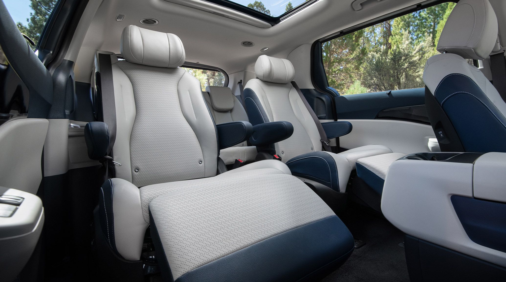 The 2027 Kia Carnival MPV Hybrid interior in gray and navy highlights its second-row VIP lounge seats fully reclined with extended leg rests, emphasizing comfort and luxury.
