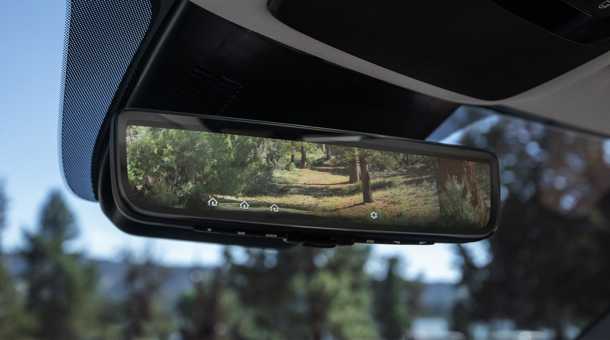 Interior view of the 2027 Kia Carnival MPV Hybrid featuring the full display digital rearview mirror, displaying a clear digital feed of the forested area behind the vehicle.
