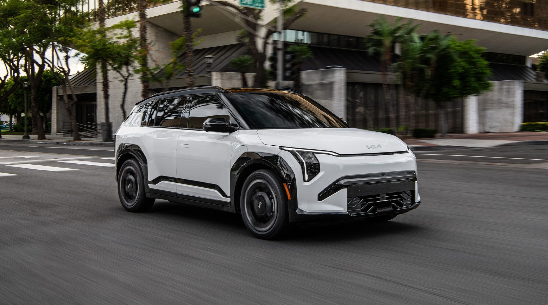 The 2027 Kia EV3 electric SUV GT-Line trim in white driving down an urban road with trees and interesting architecture.