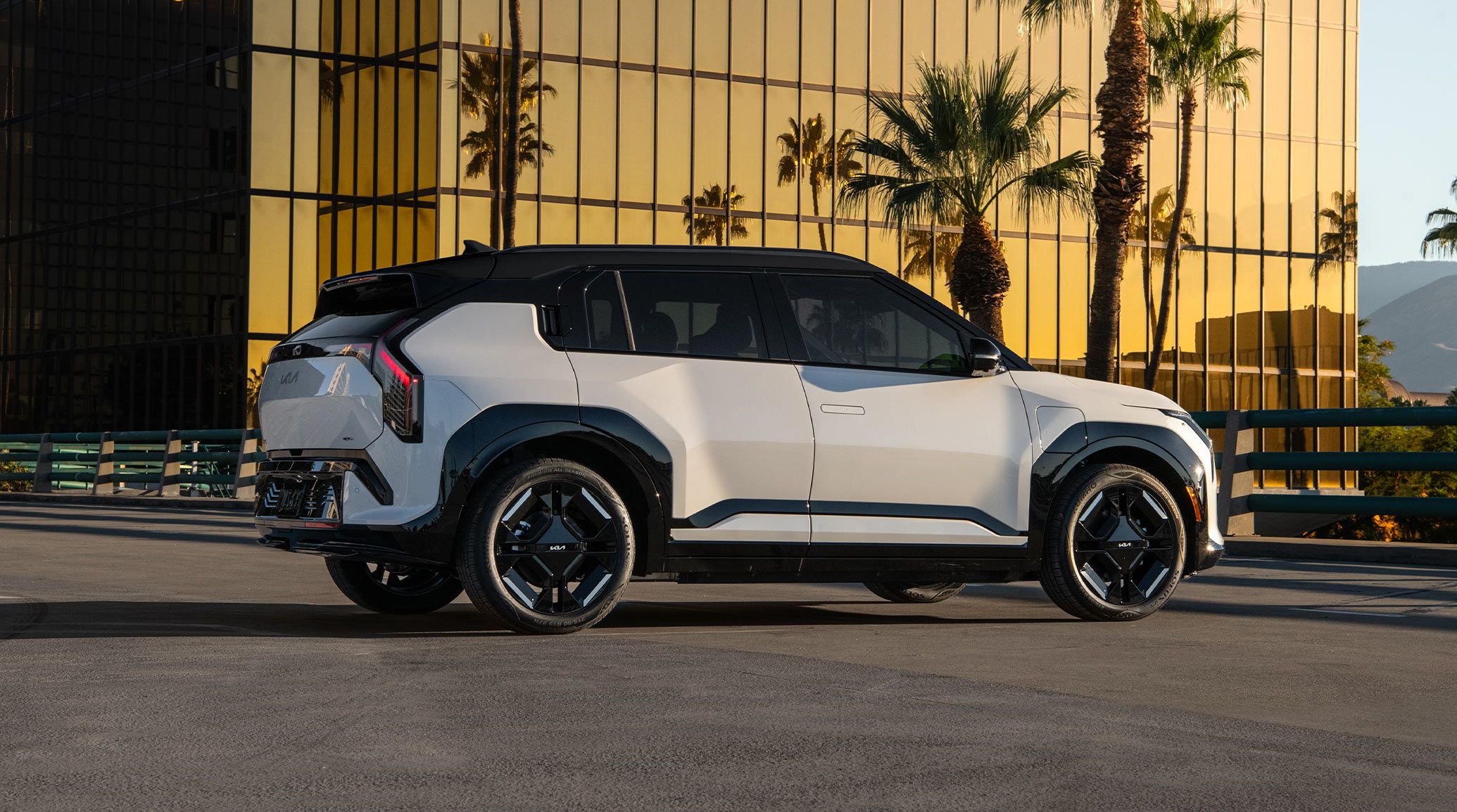 Exterior rear view of the 2027 Kia EV3 SUV parked on asphalt in an urban setting, with a glass building reflecting golden sunlight amplifying the striking silhouette of the SUV, including its rear trunk and black highlighting.