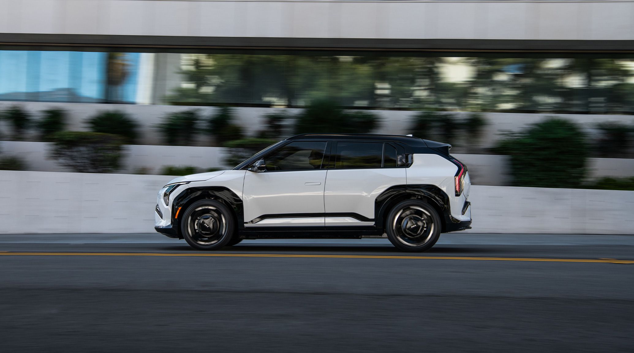 Side silhouette view of the 2027 Kia EV3 compact electric SUV in white with black highlights as it drives quickly over an urban road, with blurred building and plant details in the background. This photo shows the side profile of the EV, along with how it shines in the sunlight.