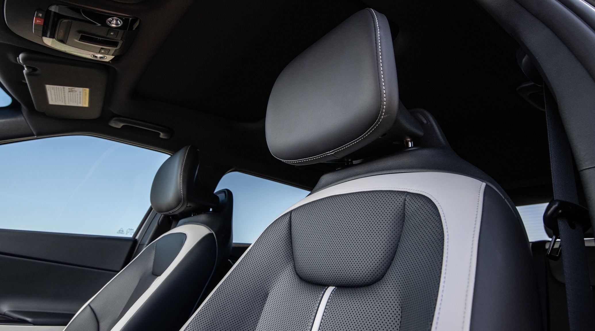 A low-angle photograph looking up at the ergonomic headrests inside the 2026 Kia EV6 electric CUV, in black and off-white SynTex seat trim, also showing the interior ceiling, coat hanger handles, seatbelts, sun visor, and other features.