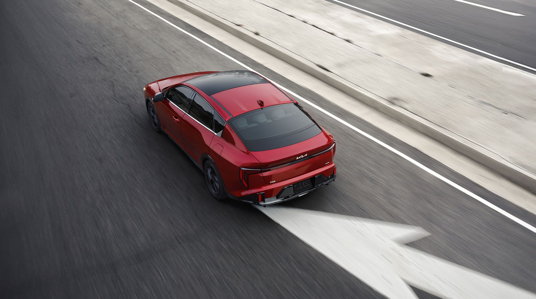 Overhead rear view of the 2027 Kia K4 sedan in red, driving down a highway in a merging traffic lane, highlighting the wide sunroof.