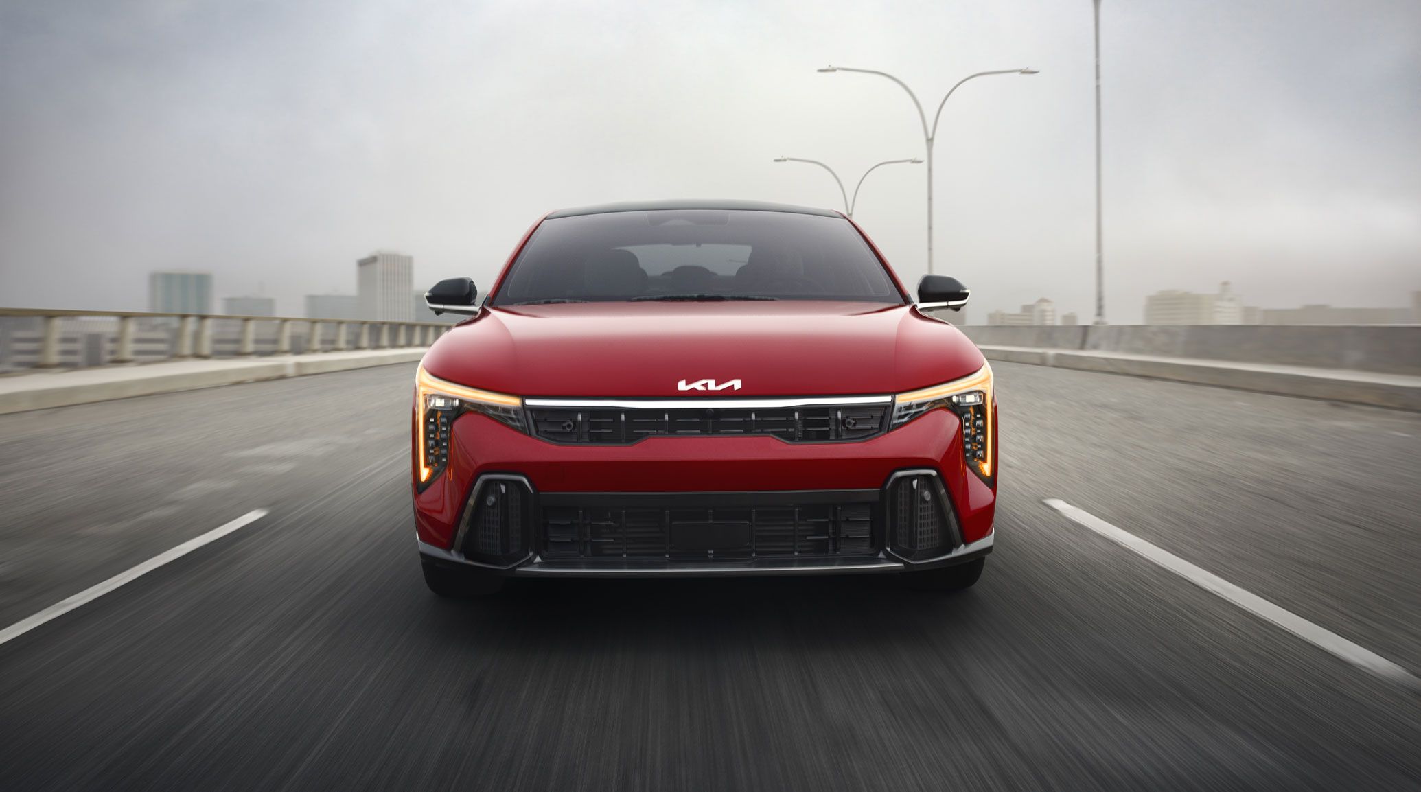 Front view of the 2027 Kia K4 sedan driving straight towards the camera, showing its windshield, hood, front grille, and amber daytime running lights on the GT-Line Turbo trim in red.