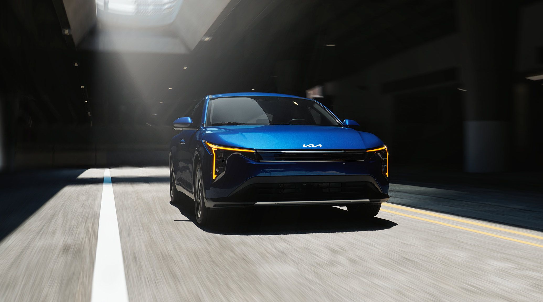 Front view of the 2027 Kia K4 EX trim in vibrant blue with dynamic light and shadows, featuring the gloss black and chrome trim grille, as it drives through an underground concrete structure.