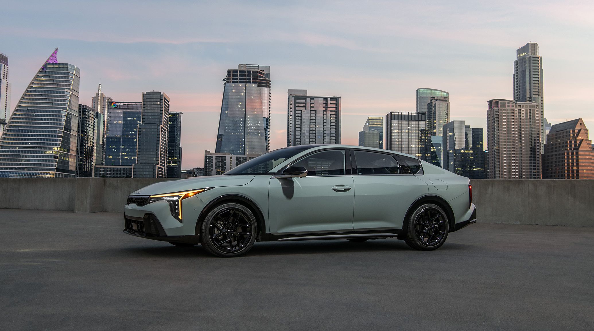 A 2027 Kia K4 shown in gray in a driver's-side view, featuring the distinct headlights and design contours as it is parked on a top level lot with city skyscrapers in the background.