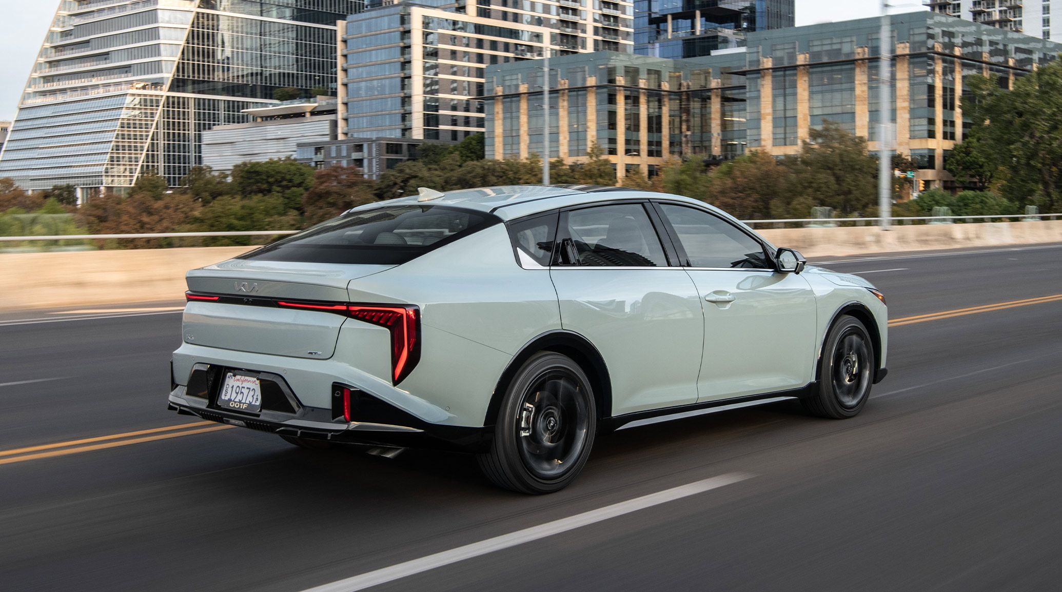 Rear three-quarter view of the 2027 Kia K4 in gray driving confidently down an empty road, with tall buildings next to the road. The car shines in the sun and features LED taillights.