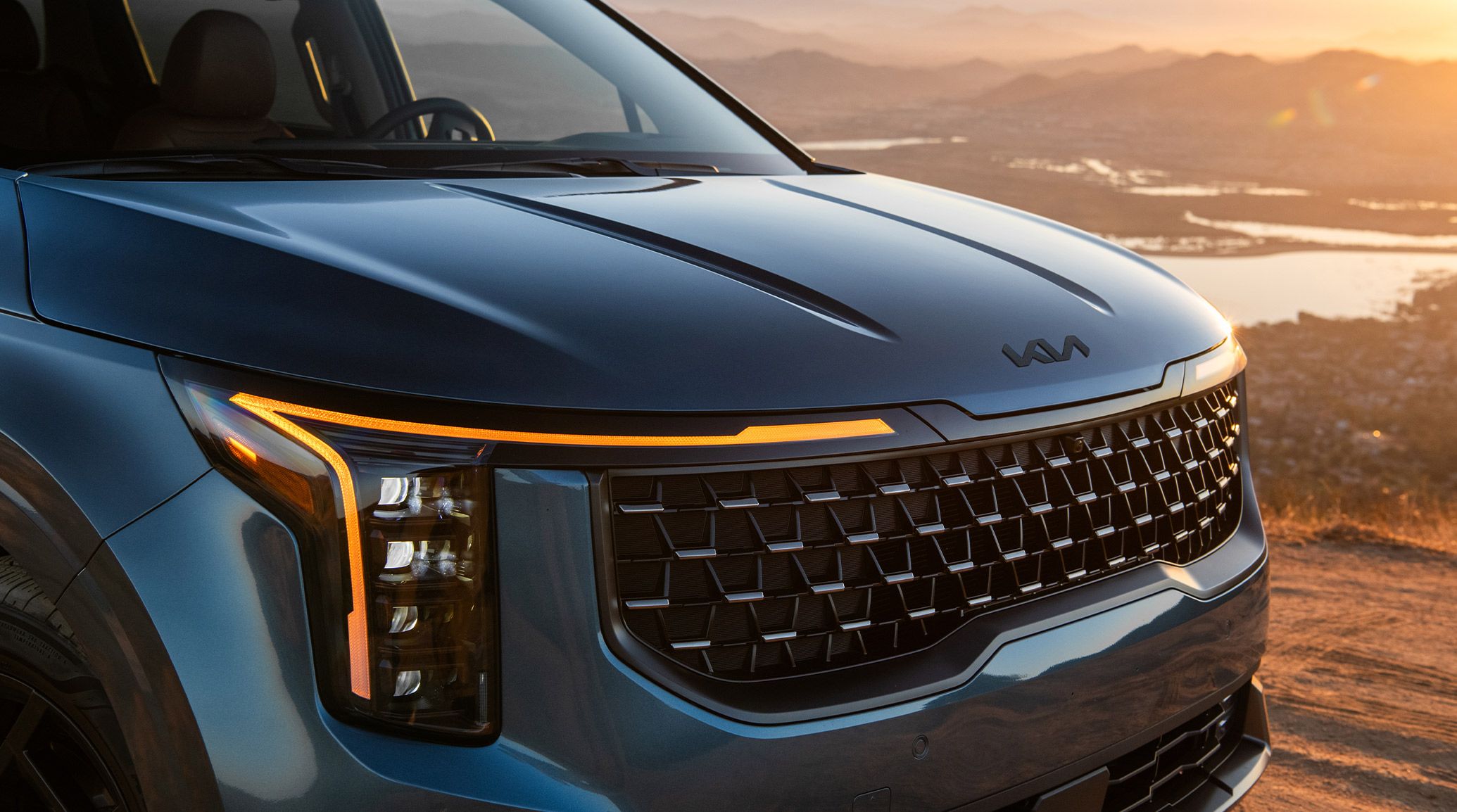 Close-up of 2027 Kia Carnival MPV SX Prestige in blue, showcasing the bold front grille and signature vertical LED headlights, set against a lake with mountains.