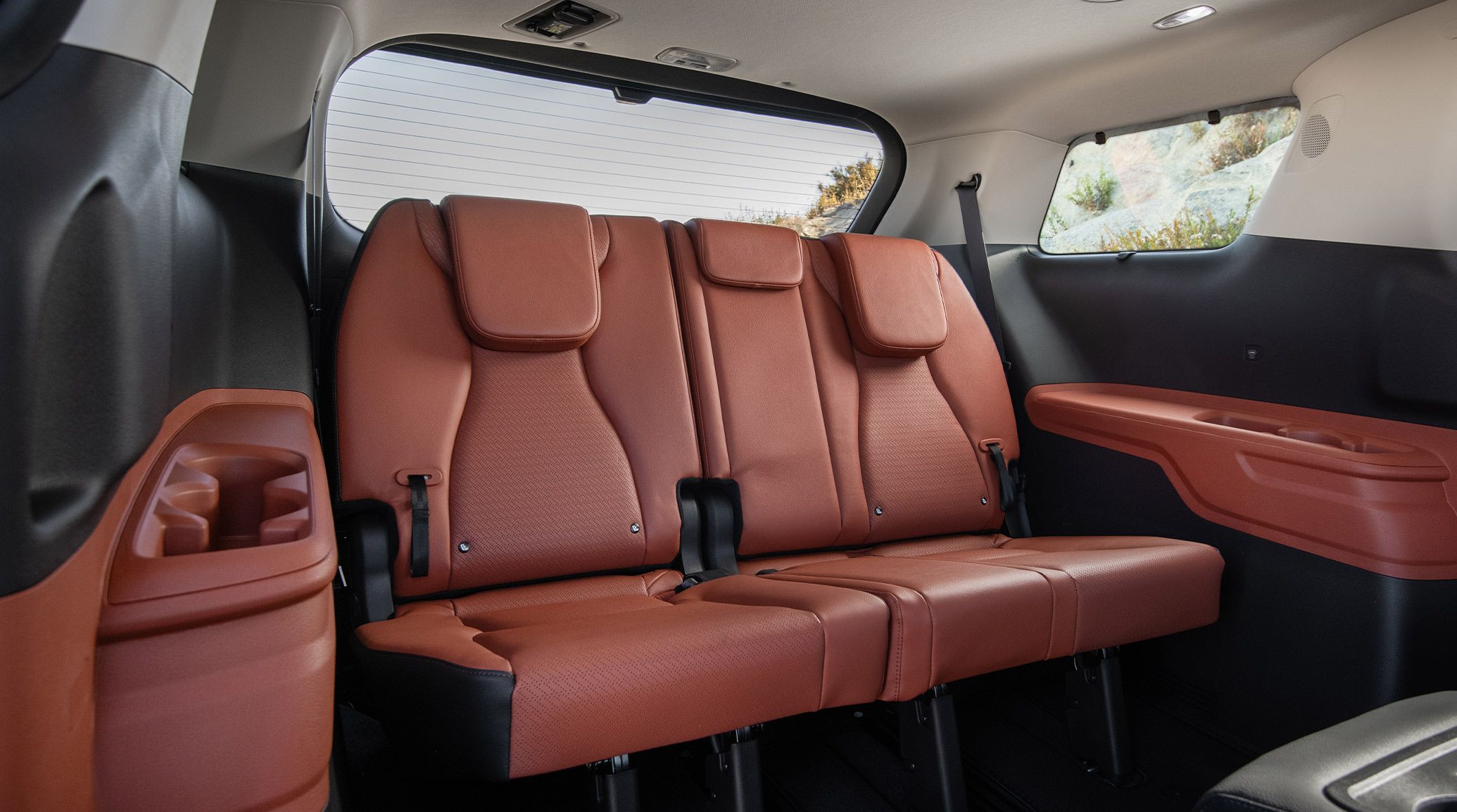 An image highlighting the red third-row seating area of the 2027 Kia Carnival MPV. The image highlights the back row seating and the spacious cupholders, with blue skies visible through the rear-view window.
