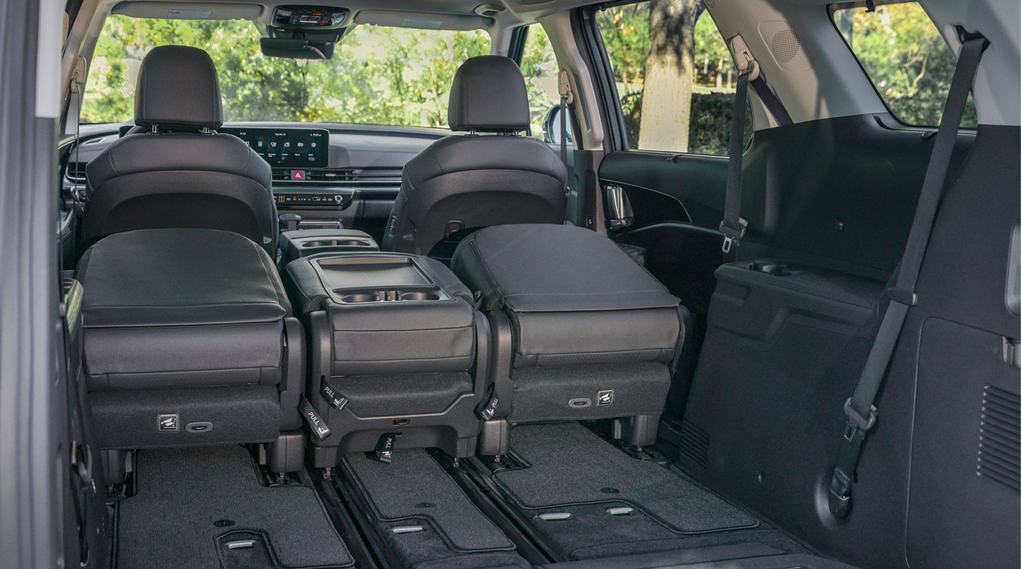 Interior view of the 2027 Kia Carnival MPV with second-row seats folded and slid forward for flexible cargo space with trees visible through the windshield.
