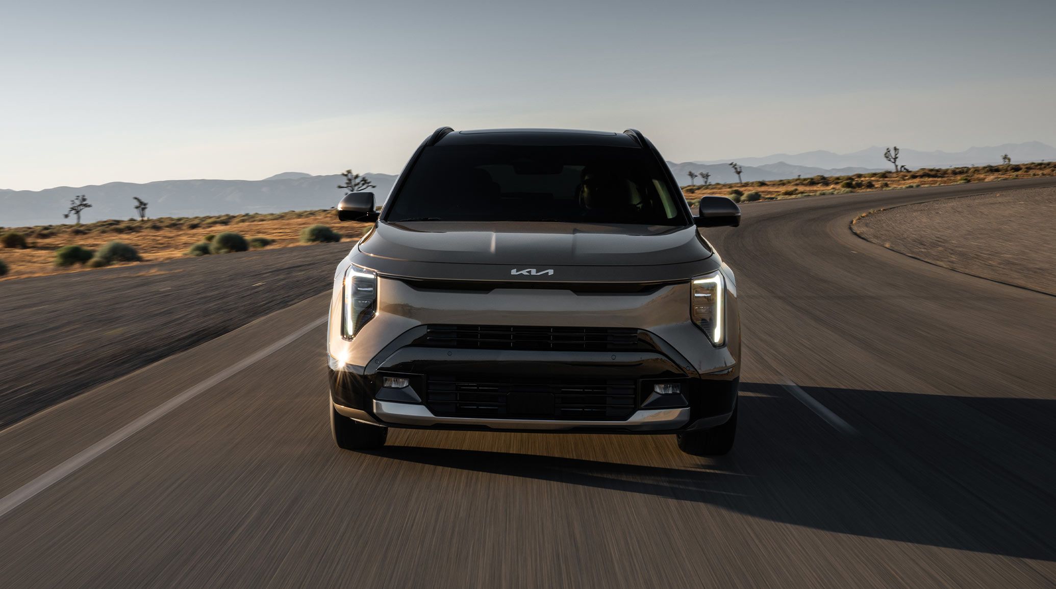 Front view of the 2027 Kia Niro Hybrid HEV sport utility vehicle as it drives around a curve on a desert road; the image highlights its LED projector headlights glowing white.