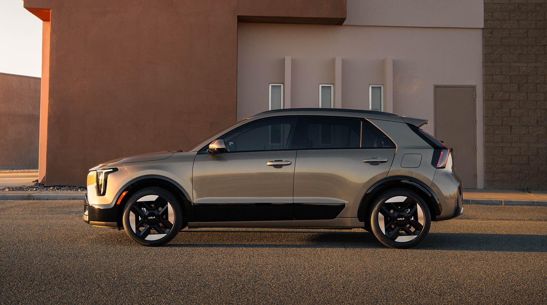 Side profile view of the 2027 Kia Niro Hybrid subcompact SUV in a bronze brown color, highlighting its distinctive shape and geometric tire designs. The EV is parked in front of a building in the desert.