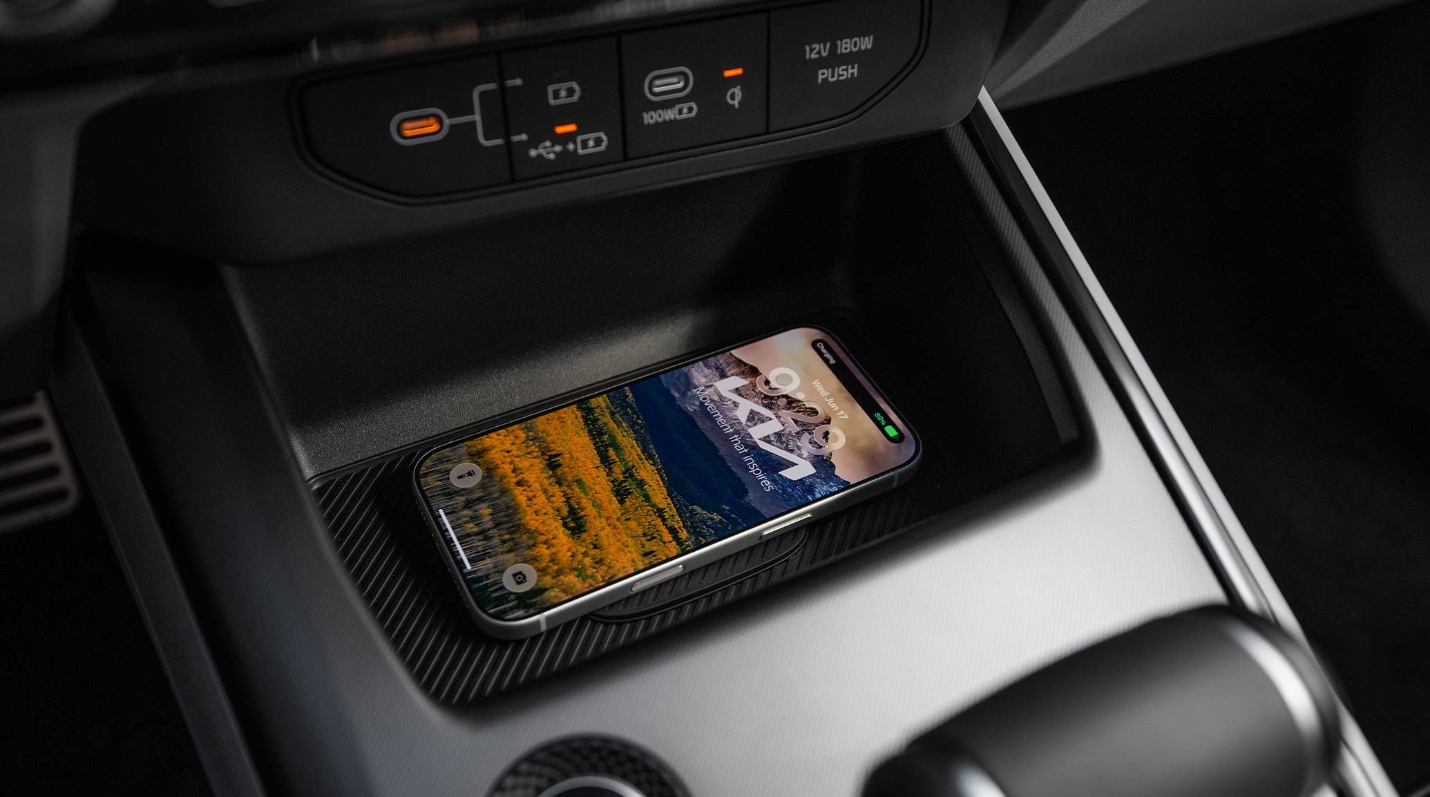 A photograph showing the front center console of a Kia vehicle, with a cell phone resting on the wireless charging surface pad. The smartphone has a landscape and Kia logo for a background. 