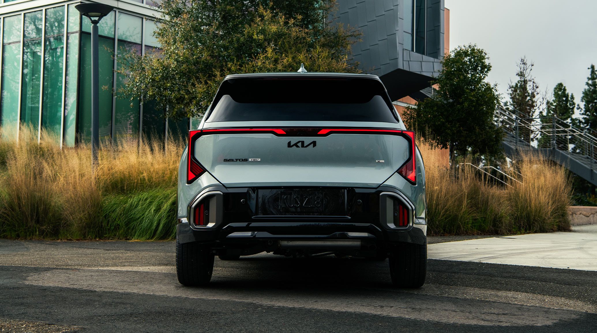 Rear shot of the 2027 Kia Seltos Hybrid in blue parked against a building with plants in front of the building, highlighting the LED taillights, sleek design, and black Kia Logo on the SUV.