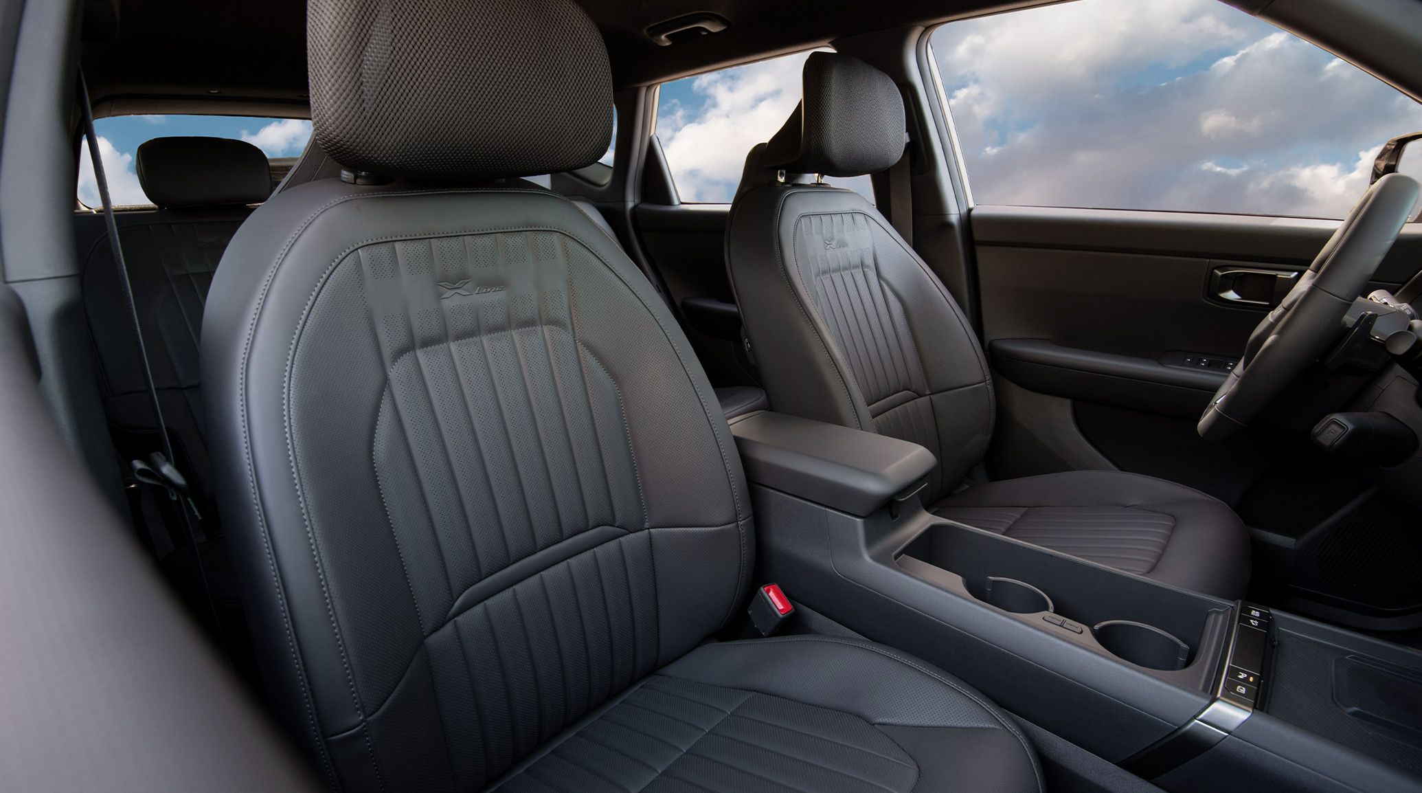 Still shot of the 2027 Kia Seltos Hybrid interior front seats featuring X-Line embossed trim and premium textured detailing, highlighting the comfortable seats and headrests. The shot also highlights the spacious cupholders, and trees are visible through the window.