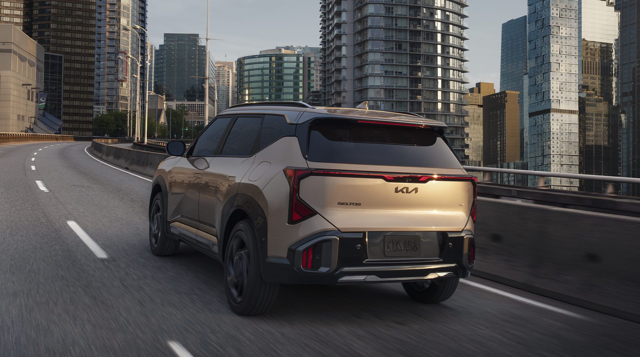 Rear shot of the 2027 Kia Seltos in brown as it drives quickly on a ramp, with an urban landscape and tall buildings around it. This photo emphasizes the SUV’s sleek trunk design and tinted windows.