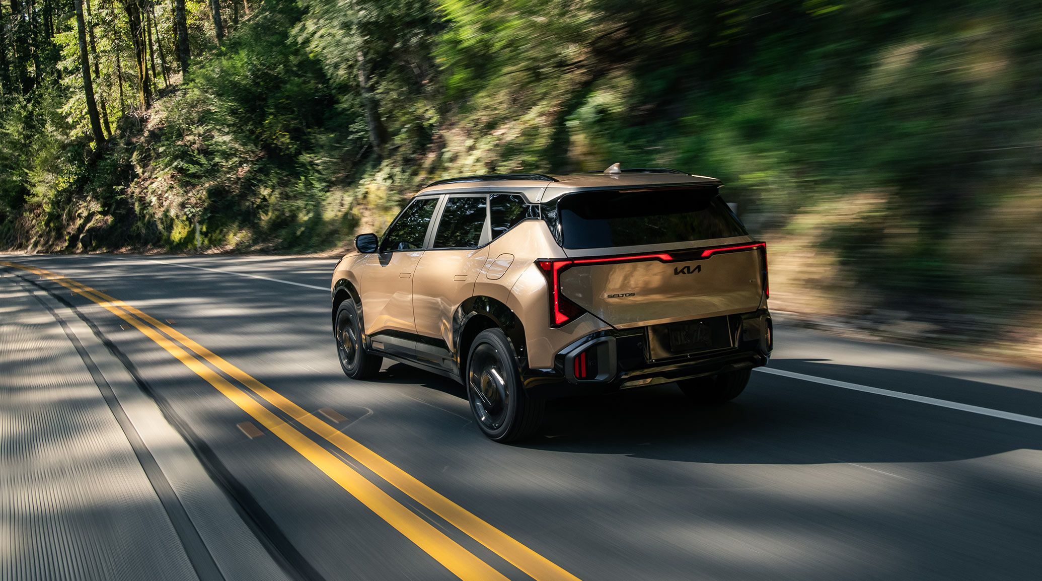 Rear side photograph of the 2027 Kia Seltos in brown as it drives quickly through a hillside forest road, with motion blur distorting the trees and rocky slope behind it. This photo emphasizes the excitement and freedom that driving a Seltos may inspire.