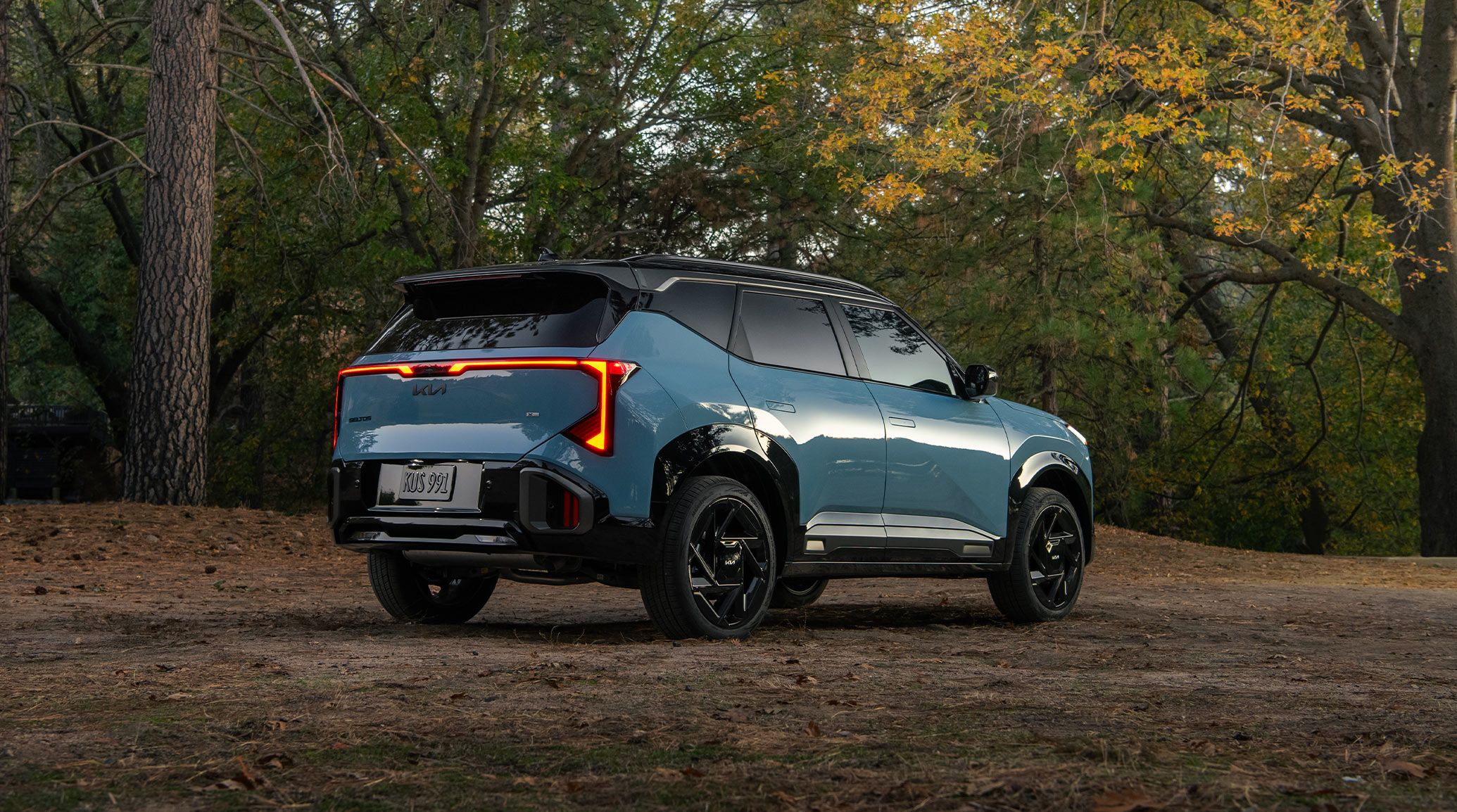2027 Kia Seltos in blue, three-quarter rear view parked on a dirt road with trees in the background, highlighting its sleek LED taillights, floating roof design, and auto-pop door handles.