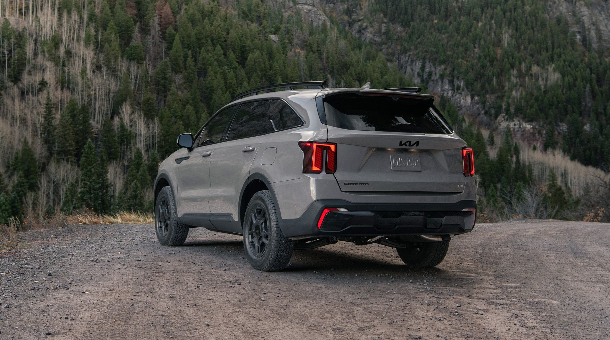 A gray 2027 Kia Sorento in a three-quarter back view parked on a dirt road right in front of a mountain filled with trees, highlighting the LED taillights and sleek trunk design.