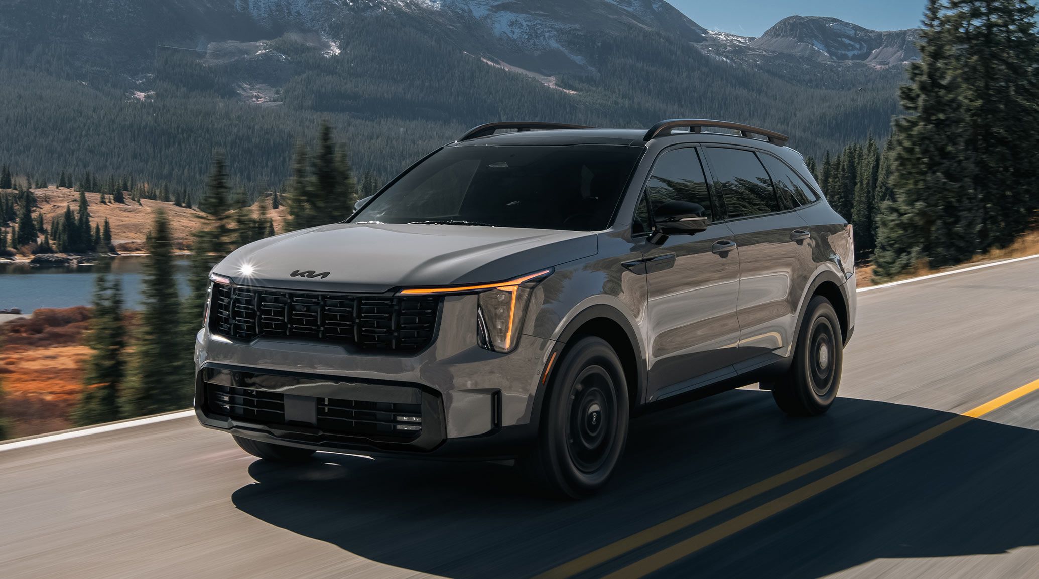 A gray 2027 Kia Sorento in a three-quarter driver's side view, highlighting the LED headlights and front grille. The SUV is driving on a road with a lake, mountains, and greenery next to it.