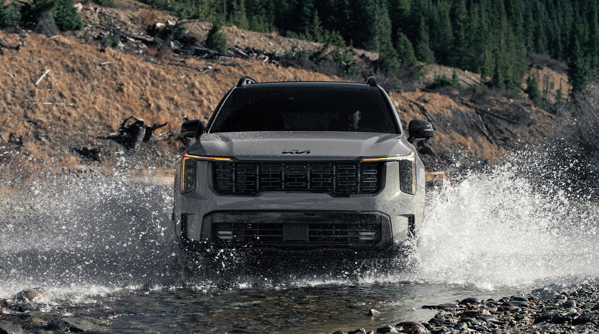 Front-view action shot of a gray 2027 Kia Sorento with trees and hills in the background. The SUV is driving through a river and is making a big splash, highlighting the all-terrain tires and unique front fascia.