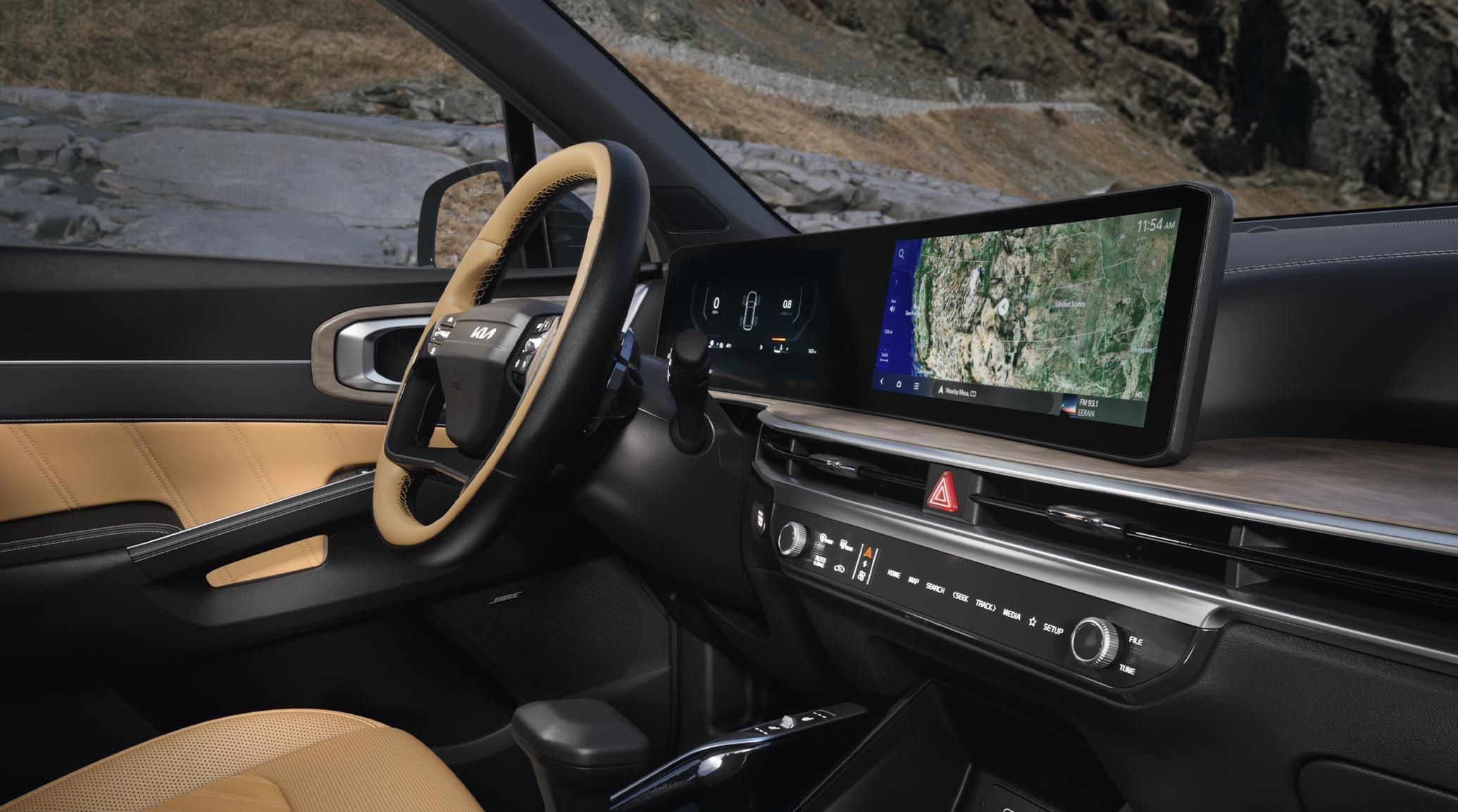 Interior side angled photo of the 2027 Kia Sorento in black and tan, showcasing its steering wheel, dual panoramic display with maps showing, center console, shifter, glove compartment, and more. Hills are visible through the driver's side window and windshield.