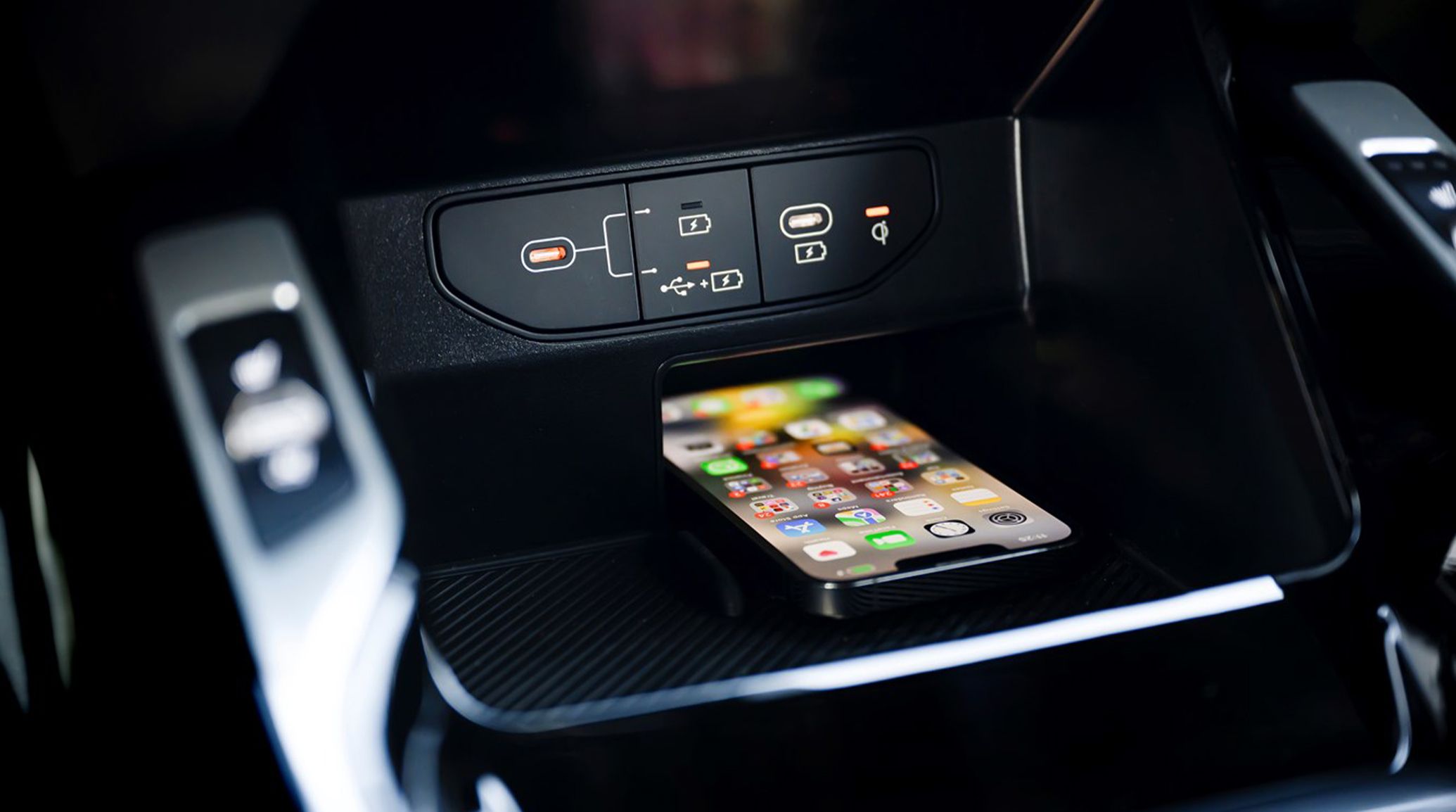 A photograph showing the front center console of a Kia vehicle, with a cell phone resting on the wireless charging surface pad. The smartphone is on the home screen, and there are 2 USB-C charging ports above it.