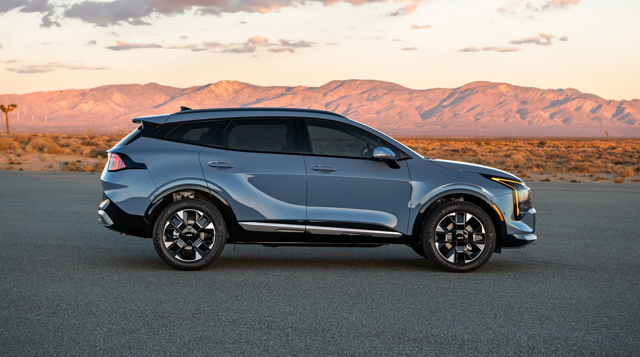 Side view of the 2027 Kia Sportage Hybrid in blue parked in the road with mountains and desert vsisble behind during sunset. The image highlights the crossover SUV's sleek design and distinctive wheels.