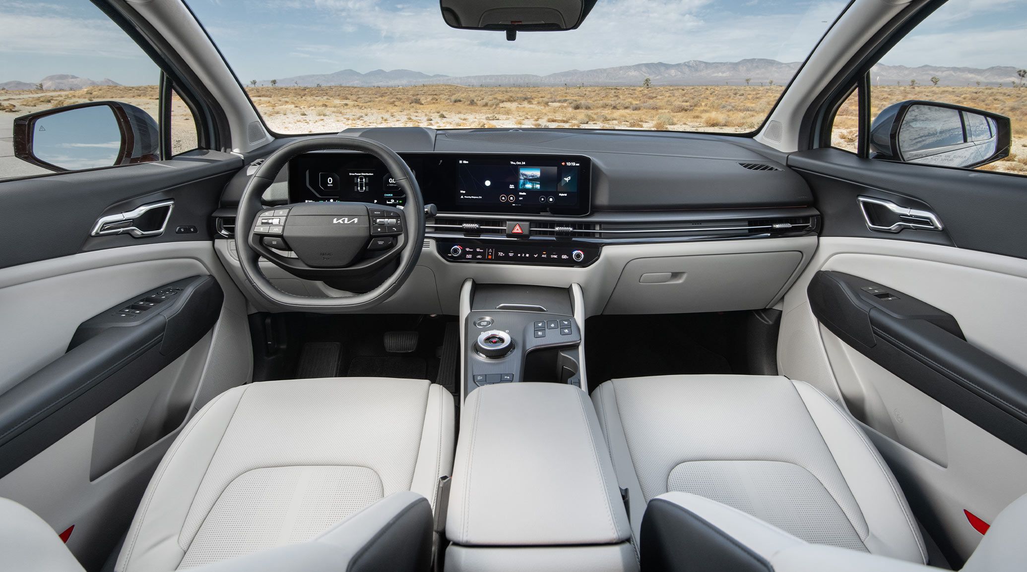 Interior view of the 2027 Kia Sportage Hybrid featuring a light gray interior with the driver-side steering wheel, digital display, sleek design, and center console highlighted, with a desert landscape and distant mountains visible through the windshield.