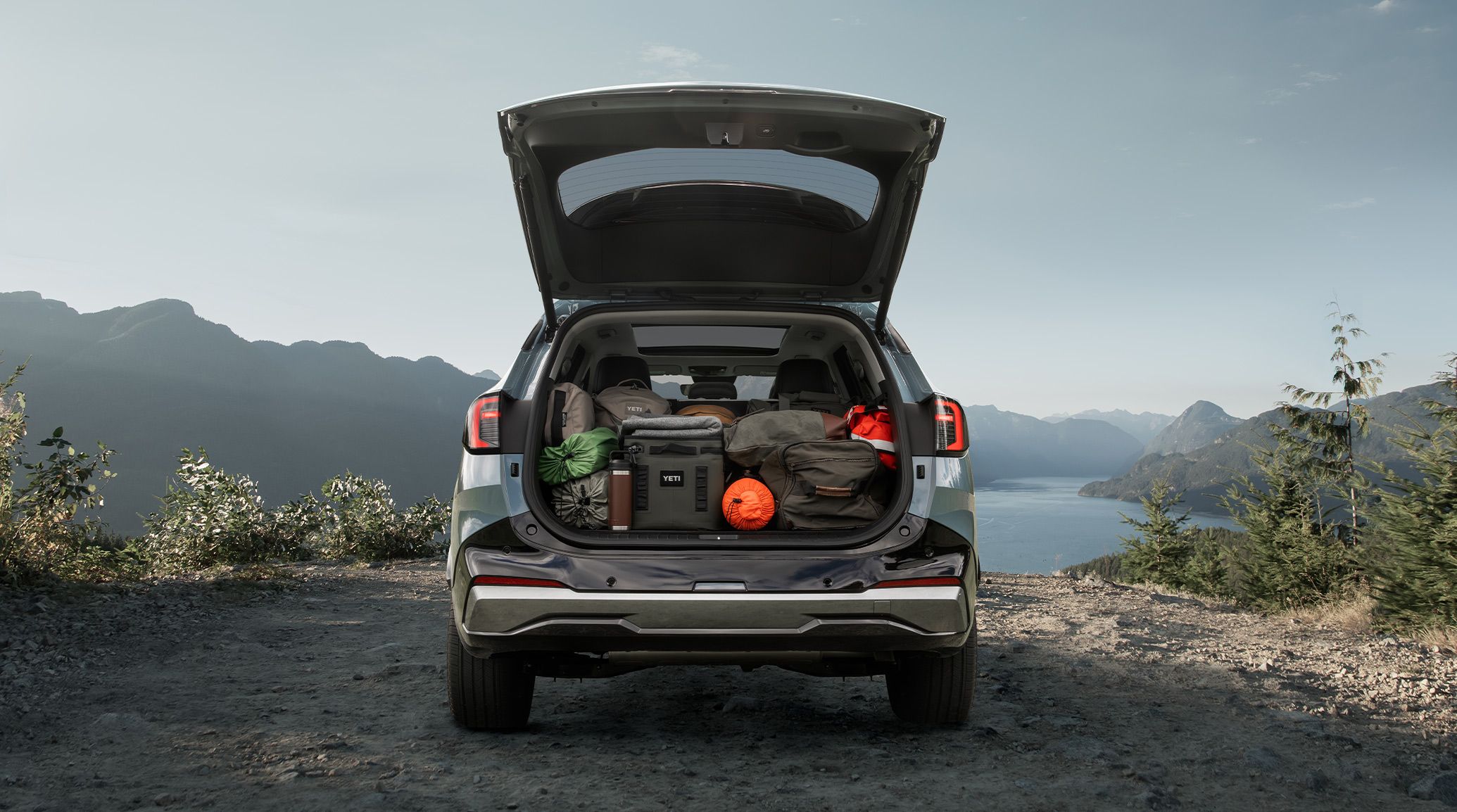 Photo of the 2027 Kia Sportage Hybrid with an open trunk showcasing the spacious cargo area packed with outdoor gear, parked set against a cliff with mountains and a lake visible.