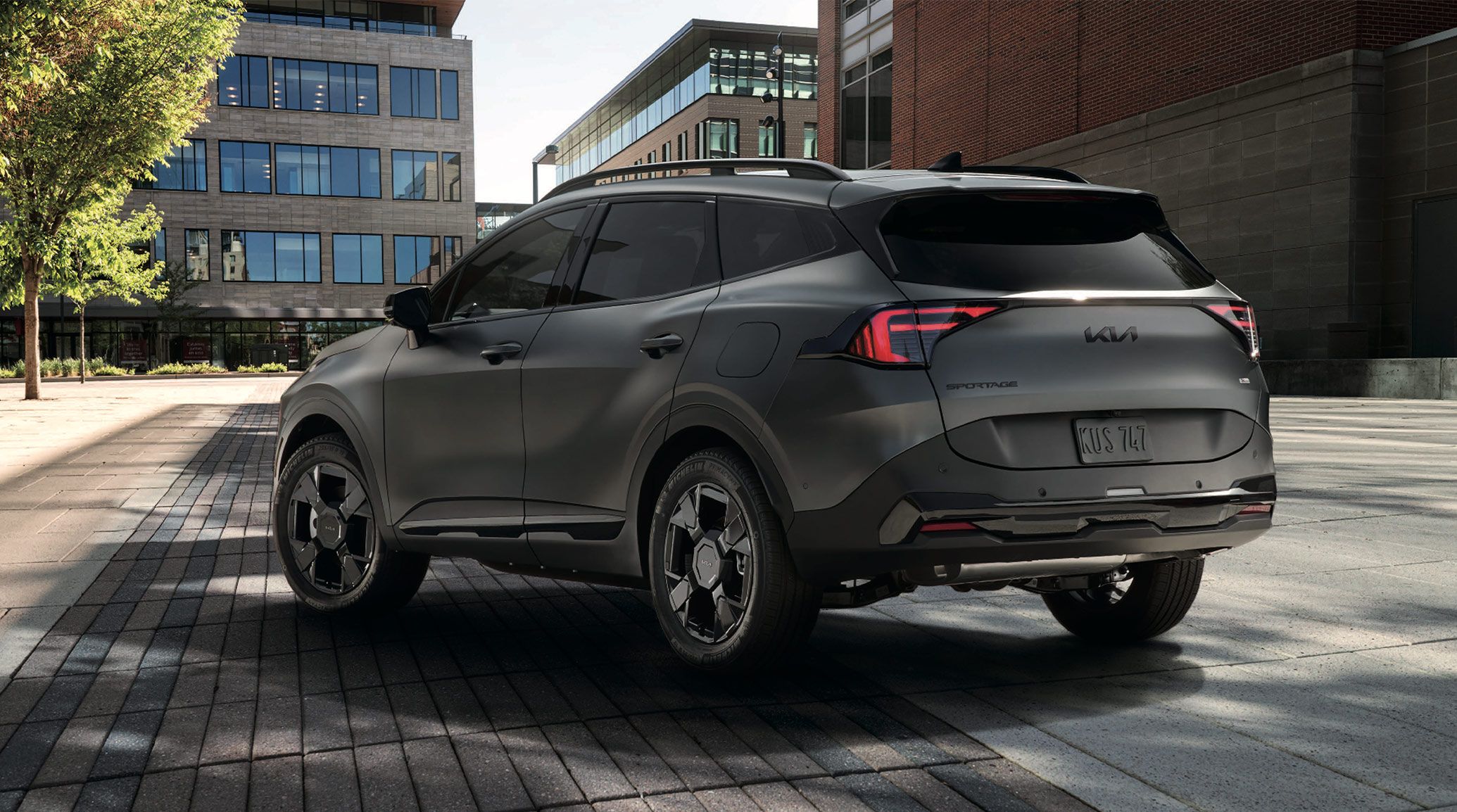 2027 Kia Sportage Plug‑In Hybrid in matte gray, viewed from the rear three‑quarter angle with distinctive taillights, sharp body lines, and city buildings surrounding the parked vehicle.