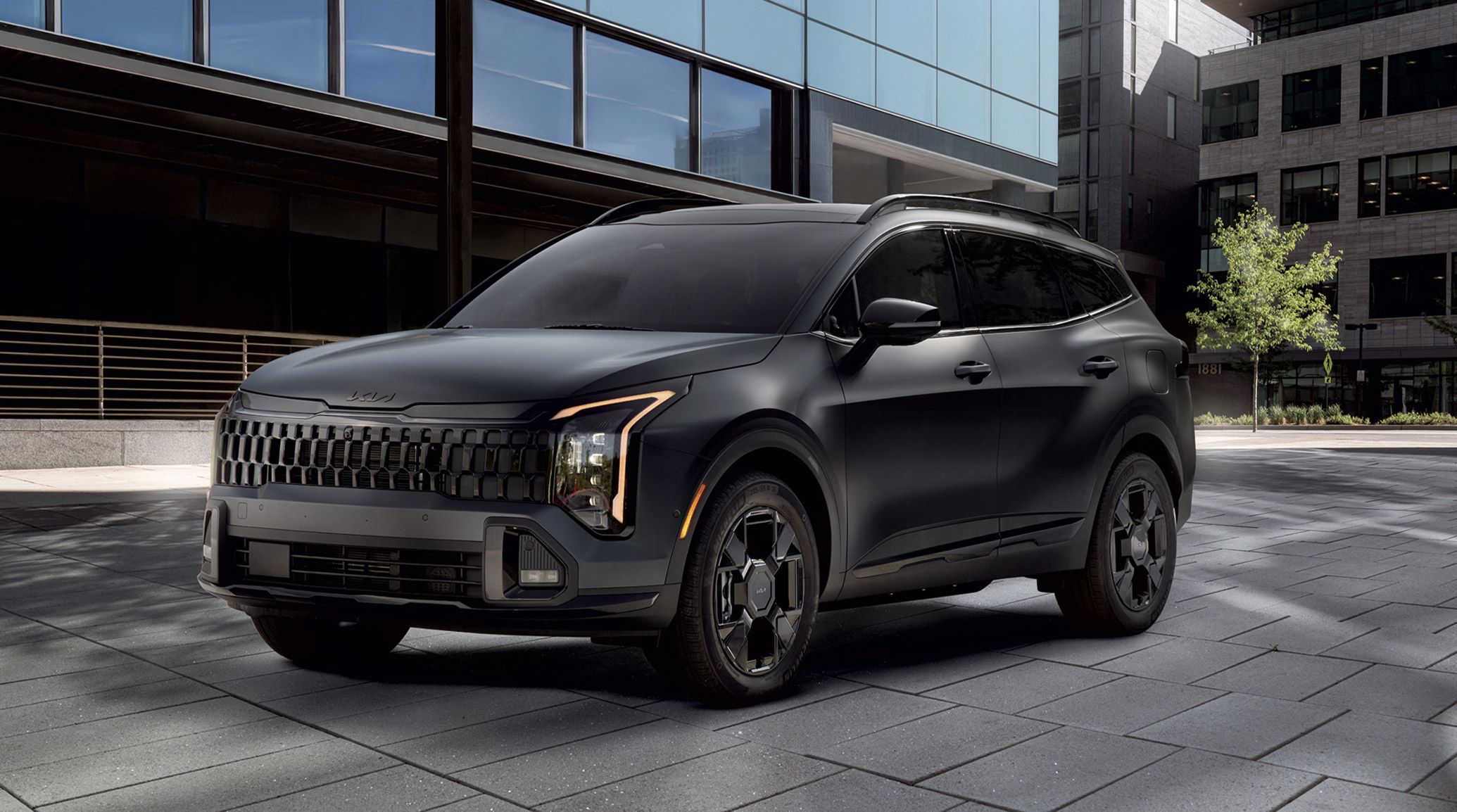 2027 Kia Sportage Plug‑In Hybrid parked in an urban setting, shown from the front with vertical LED headlights, a bold grille design, and modern architectural buildings in the background.